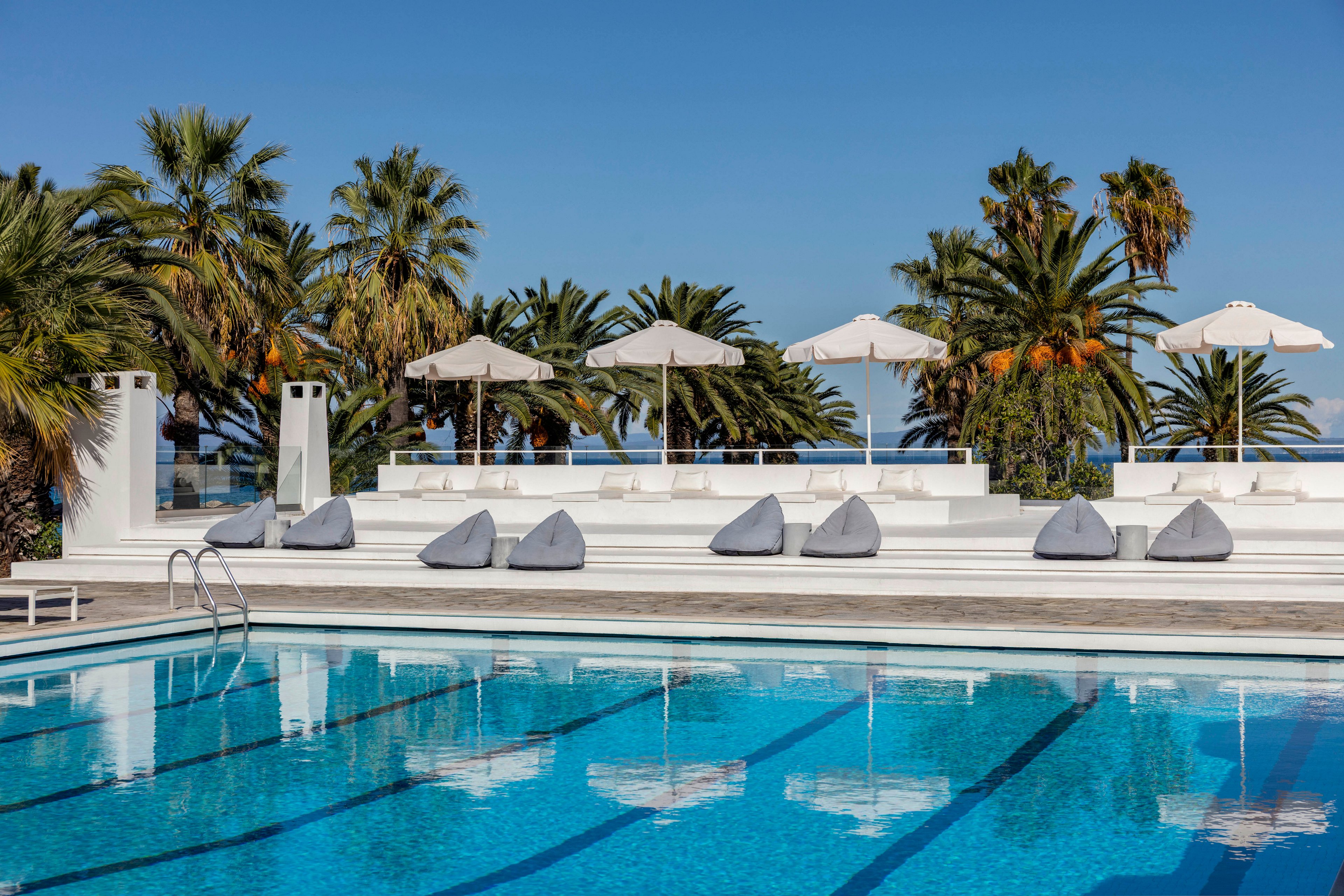 A clear blue swimming pool sits in front of a modern white terrace at a resort. Gray beanbag loungers line the deck beneath white sun umbrellas, creating a relaxed vacation atmosphere. Tall palm trees rise behind the terrace against a bright blue sky, with the sea visible in the distance. The scene suggests a sunny, tranquil day at a coastal hotel or spa.