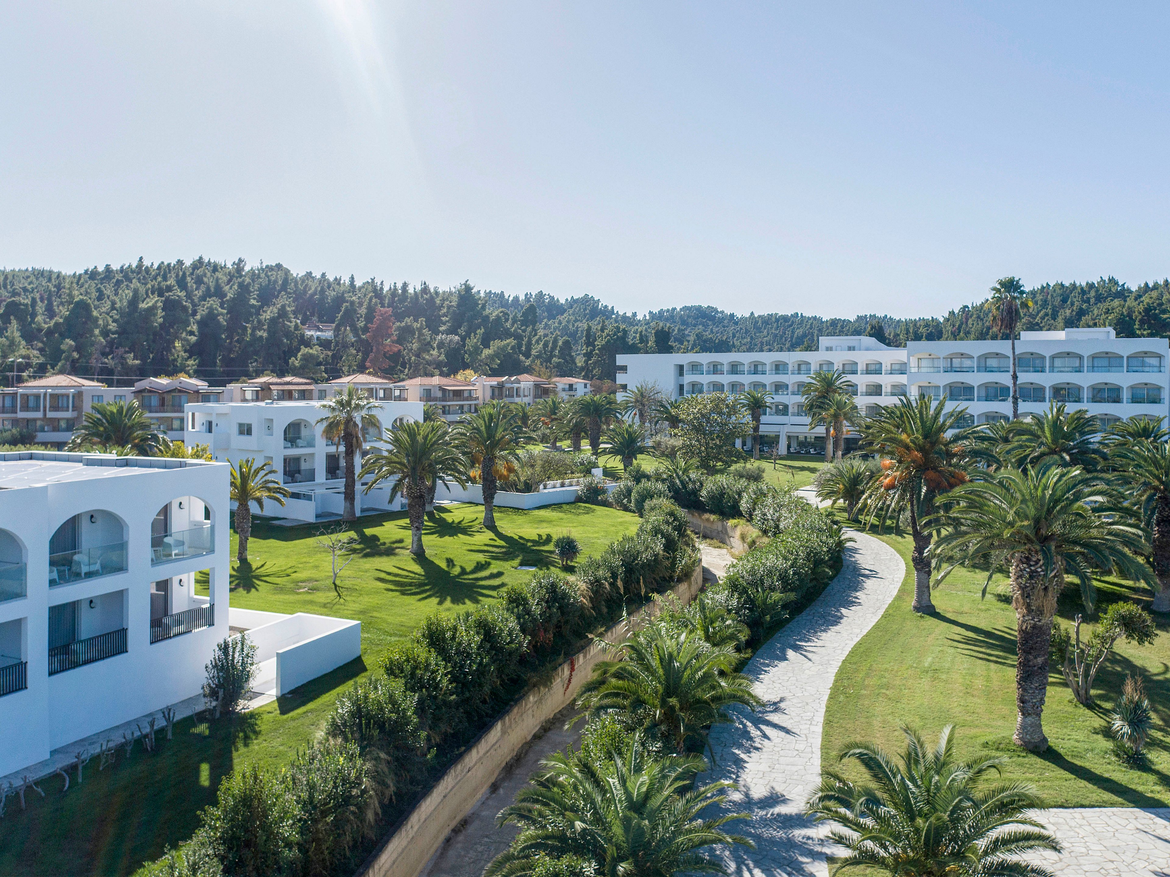 A large coastal resort complex features modern white buildings surrounded by lush greenery. Palm trees line a winding stone pathway that curves through manicured lawns and landscaped gardens. The architecture includes arched windows and balconies, giving the property a Mediterranean or tropical feel. Forested hills rise in the background under a clear blue sky, suggesting a sunny vacation destination.
