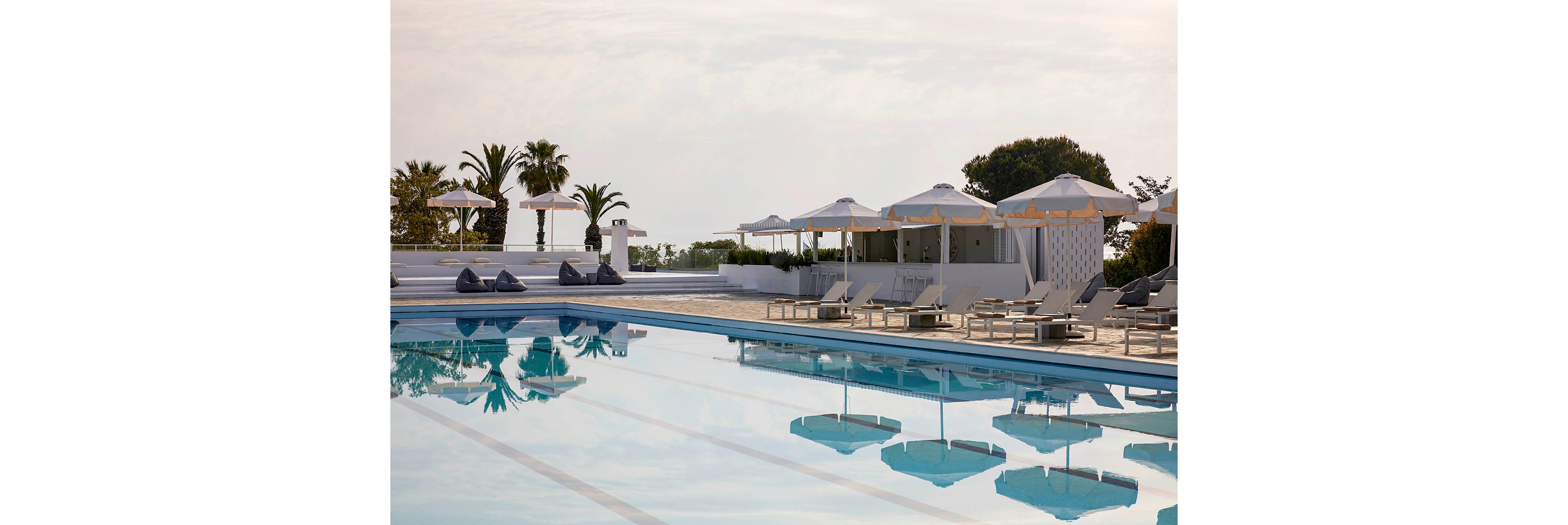 An outdoor swimming pool reflects white sun umbrellas and lounge chairs in a tranquil resort setting. Palm trees and greenery frame the pool area, suggesting a warm coastal or Mediterranean location. The scene appears calm and quiet, with soft daylight and muted colors creating a relaxed atmosphere. Cushioned seating and cabana-style structures add to the sense of comfort and luxury.