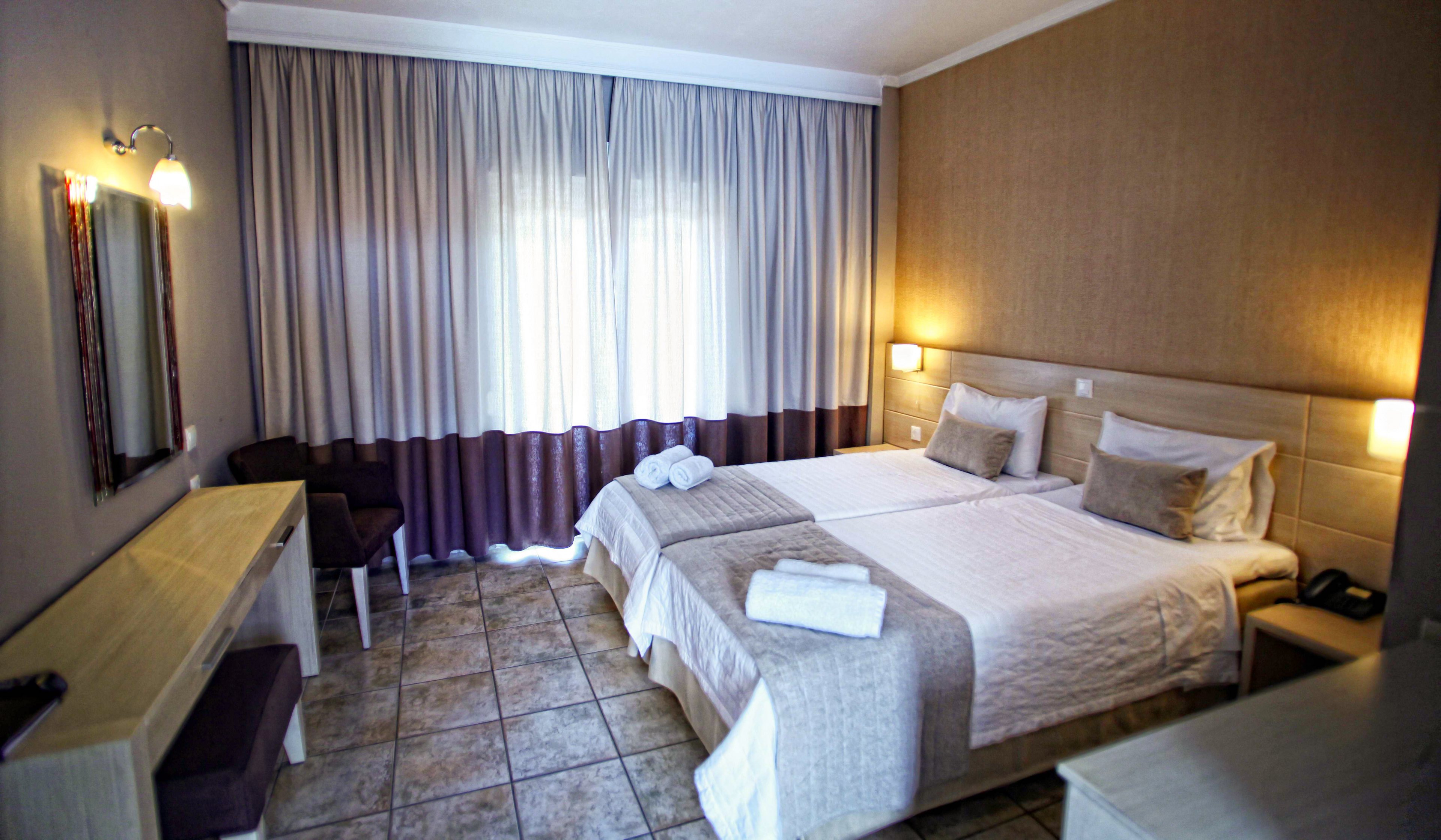 A neatly arranged hotel room features two twin beds pushed together with white linens and folded towels on top. The space includes a long desk with a mirror, chair, and bedside tables with lamps. Soft curtains cover a large window, allowing diffused daylight to brighten the room. Neutral tones and simple decor create a calm, comfortable atmosphere suitable for travelers.