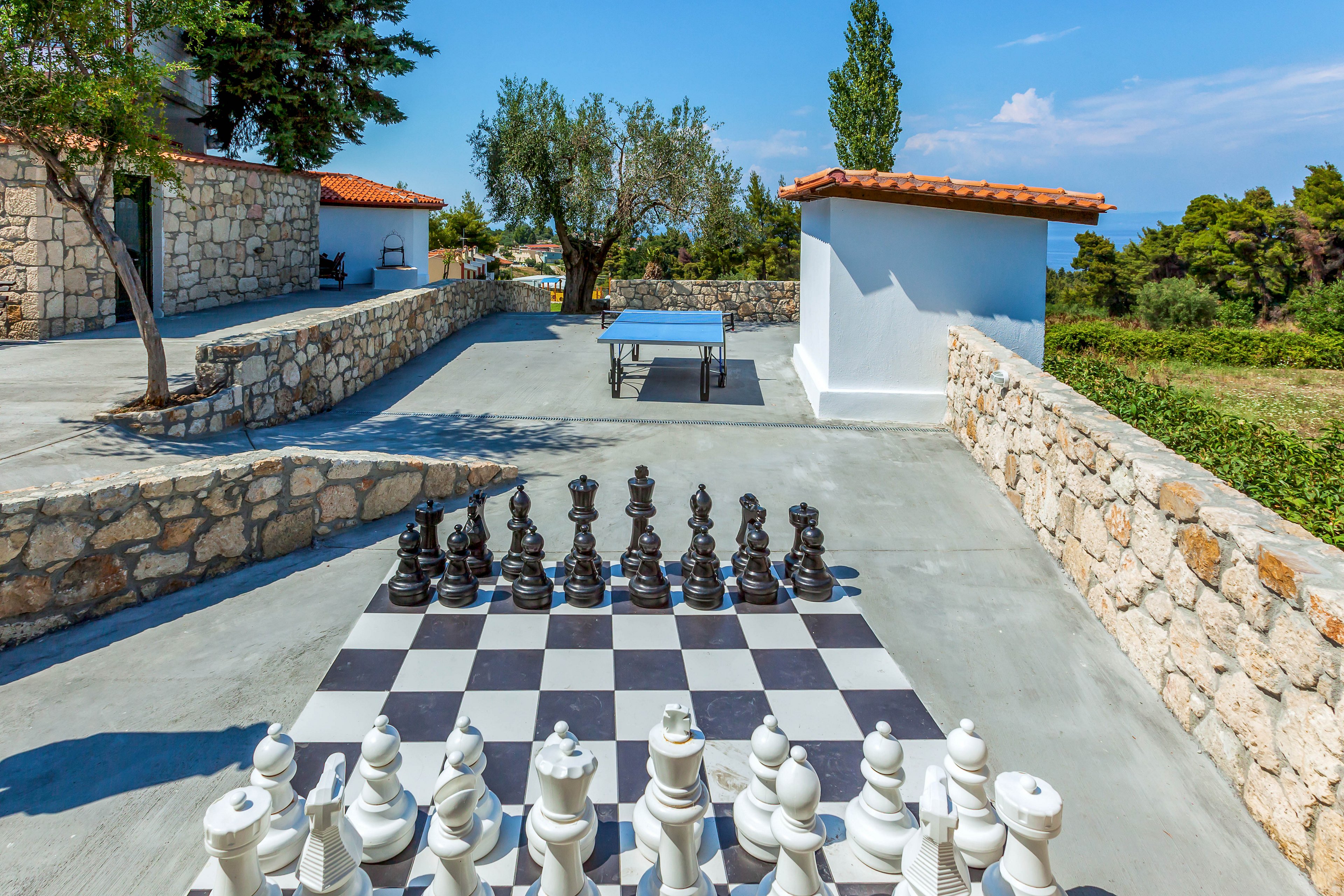 A large outdoor chessboard with oversized black and white pieces is set up on a sunny terrace. The scene is surrounded by stone walls and white buildings with red-tiled roofs. A ping pong table stands in the background, adding to the recreational atmosphere. Trees and distant greenery frame the space, with a glimpse of blue sky and sea on the horizon.