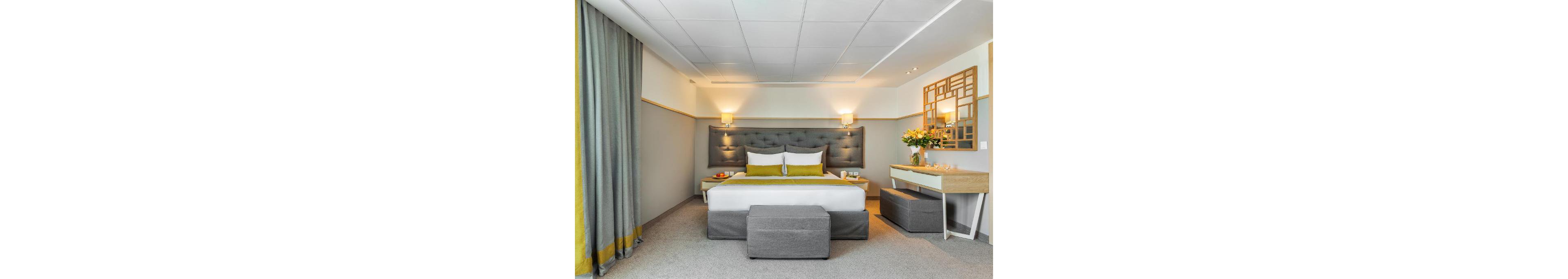 A contemporary hotel bedroom features a large bed with a tufted headboard and coordinated pillows. The room is decorated in soft gray and yellow tones, with matching curtains and upholstered furniture. Wall-mounted lamps provide warm lighting above bedside tables holding drinks and accessories. A desk with a mirror and a vase of flowers adds a touch of elegance, while clean lines and minimal decor create a calm, inviting atmosphere.