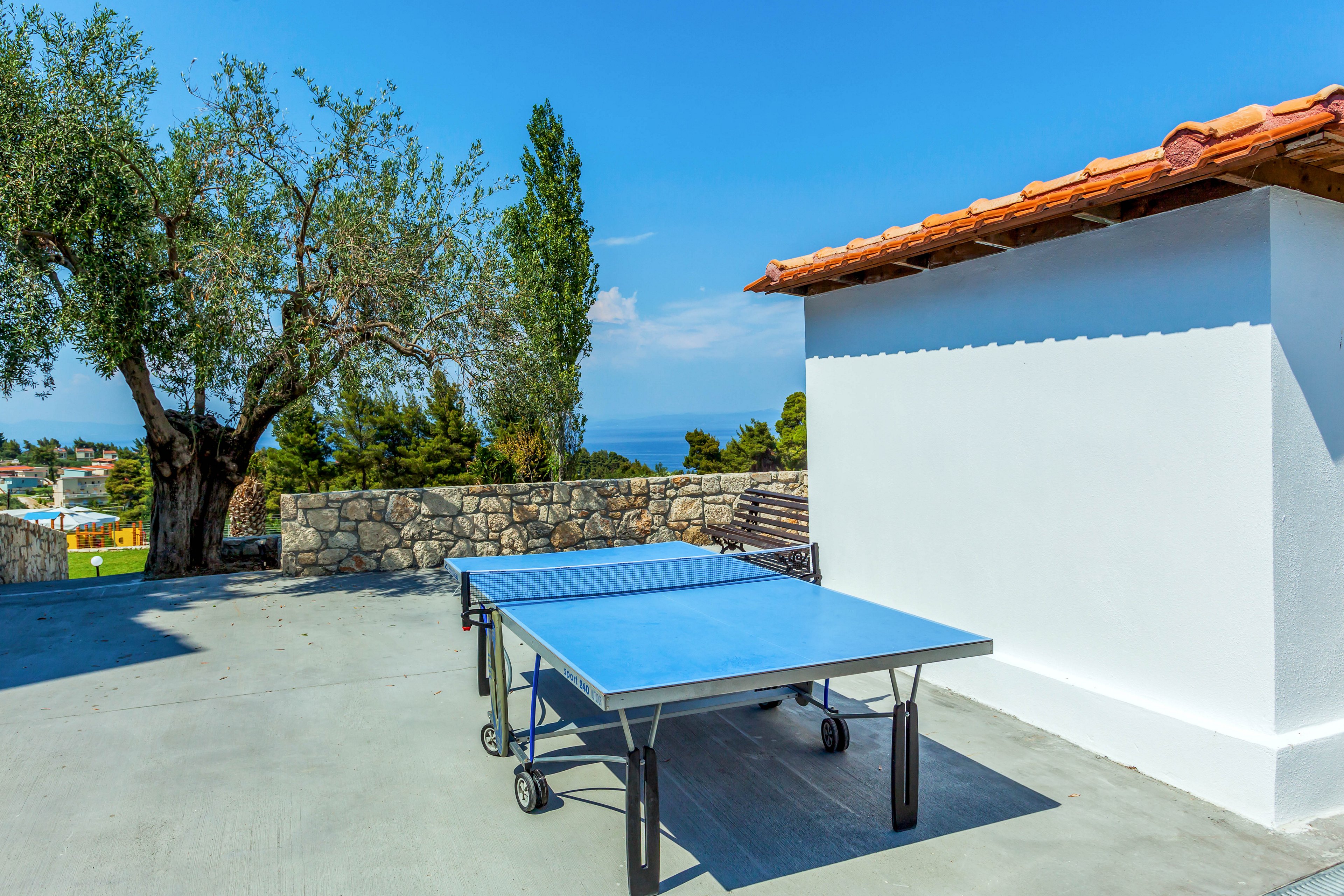 A blue ping pong table stands on a spacious concrete terrace under a clear blue sky. The terrace is bordered by a stone wall and overlooks a distant sea view. A large tree provides natural greenery next to a white building with a red-tiled roof. The scene suggests a relaxed outdoor recreation area in a warm, coastal setting.