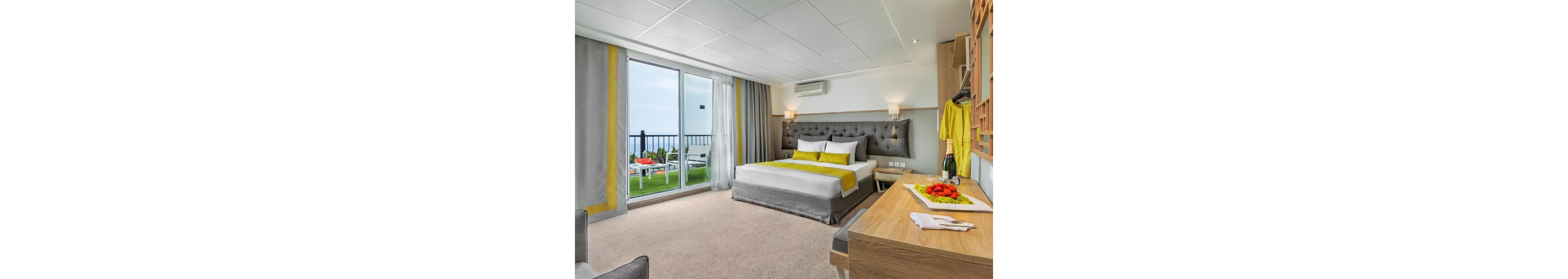 Contemporary hotel room features a large double bed with gray and yellow accents. A sliding glass door opens to a balcony with outdoor seating and an ocean view. Inside, a wooden desk, wall-mounted TV, and open wardrobe add functional comfort. Fresh fruit and a bottle of wine on the desk enhance the welcoming, vacation atmosphere.