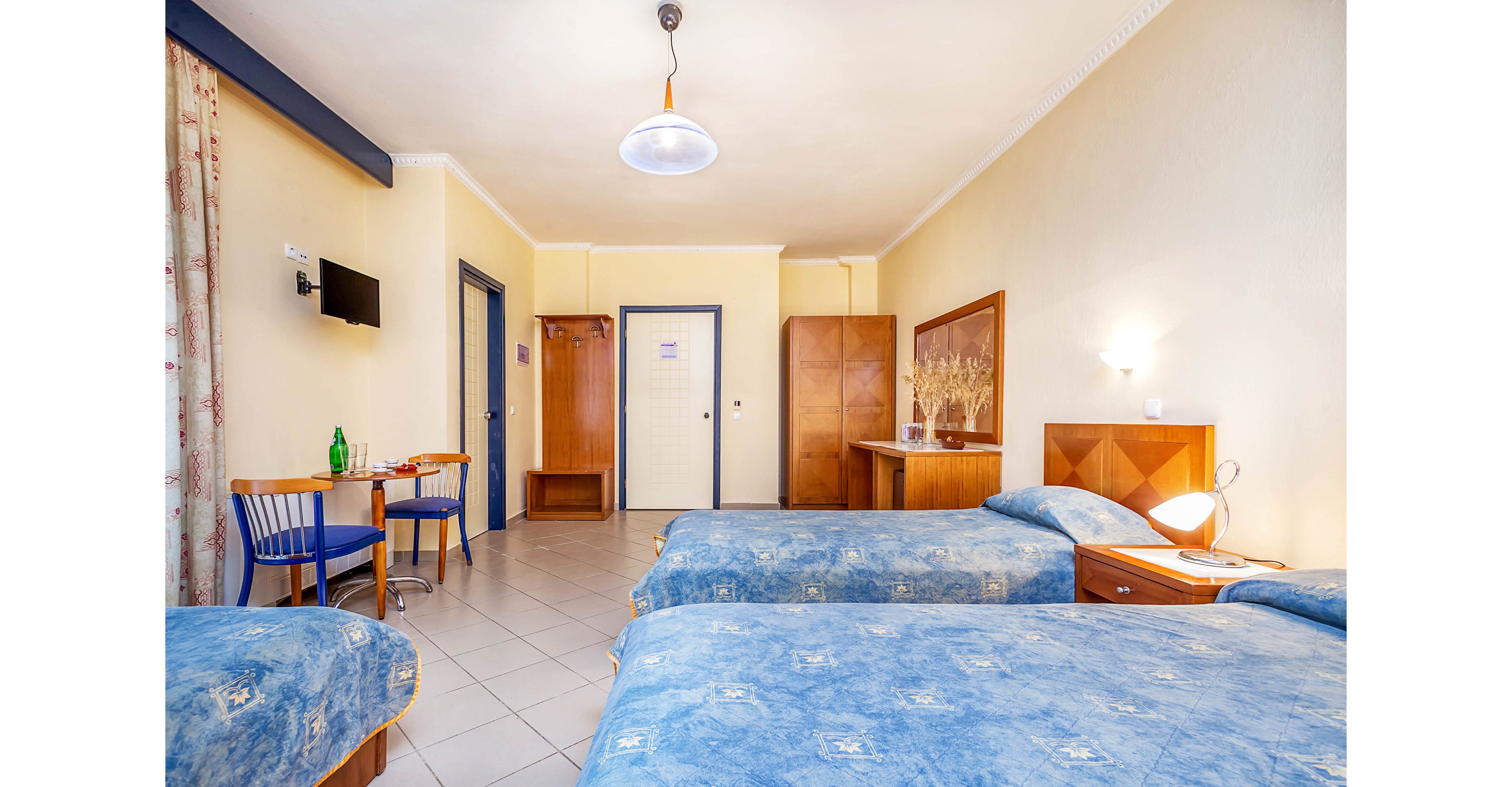Spacious hotel room featuring two blue twin beds with wooden headboards. The room includes a small wooden table with two chairs, a wall-mounted television, and a wardrobe with additional storage. Neutral cream walls and tiled flooring create a clean, simple atmosphere. Soft lighting from wall lamps and a ceiling light adds warmth to the interior.