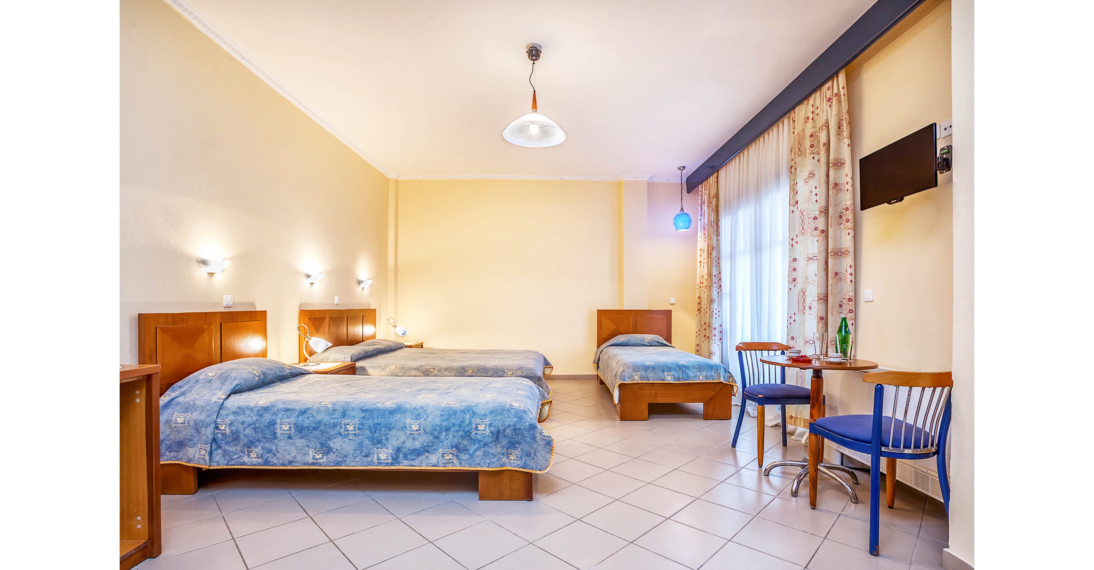Spacious hotel room featuring three neatly made single beds with blue patterned bedspreads. The room has light-colored tiled flooring, pale yellow walls, and wooden headboards with wall-mounted reading lights. A small round table with two blue chairs sits near a window or balcony door with floral curtains. A flat-screen television is mounted on the wall, and a bottle with glasses is placed on the table, suggesting a comfortable stay for multiple guests.