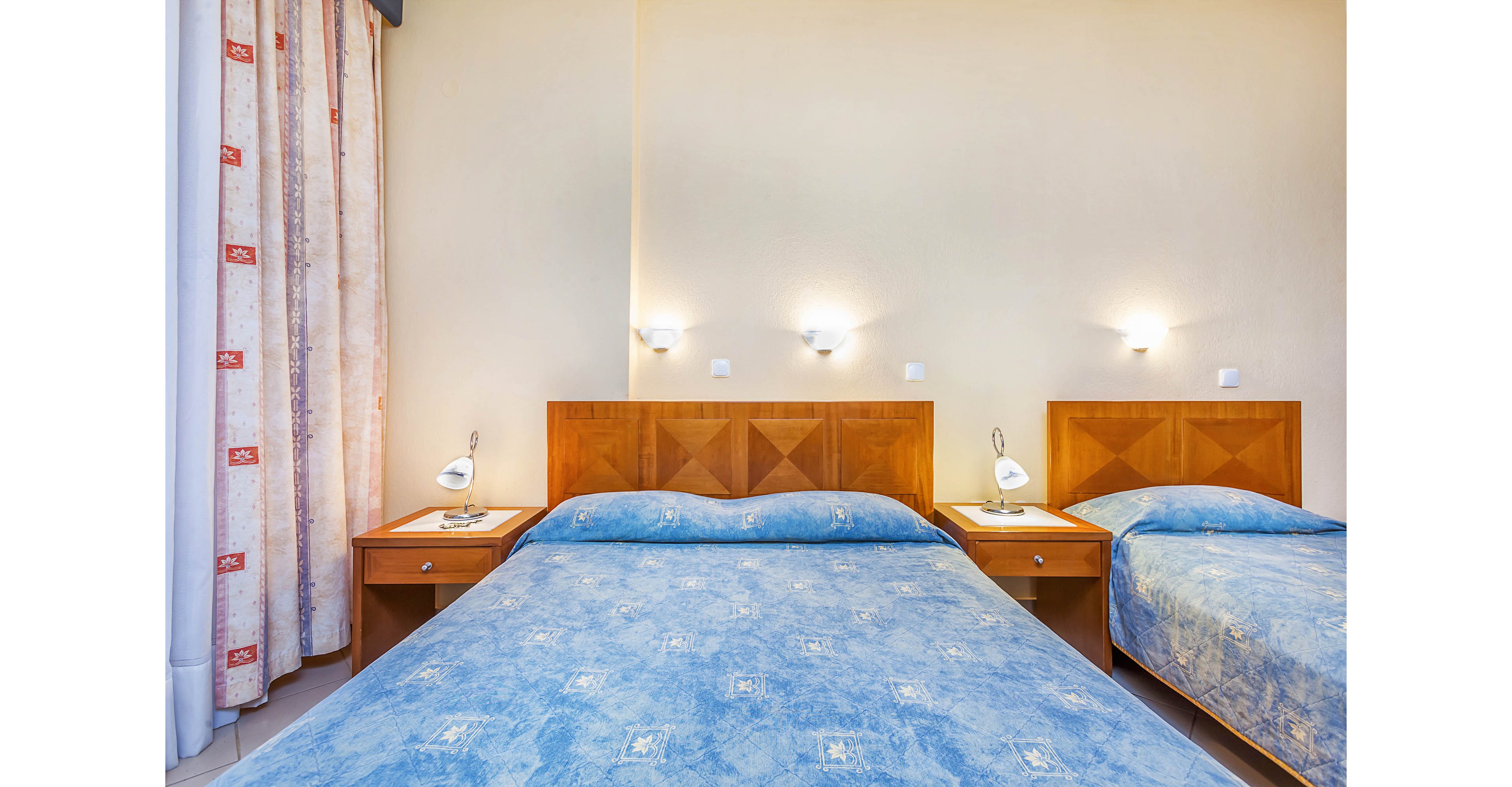 Interior view of a tidy hotel room featuring a double bed and a single bed with matching blue bedspreads. The beds have wooden headboards and are flanked by small bedside tables with reading lamps. Wall-mounted lights cast a warm glow on the pale yellow walls, creating a clean and inviting atmosphere. Light-colored curtains with a subtle pattern hang to the side, adding softness to the simple decor.
