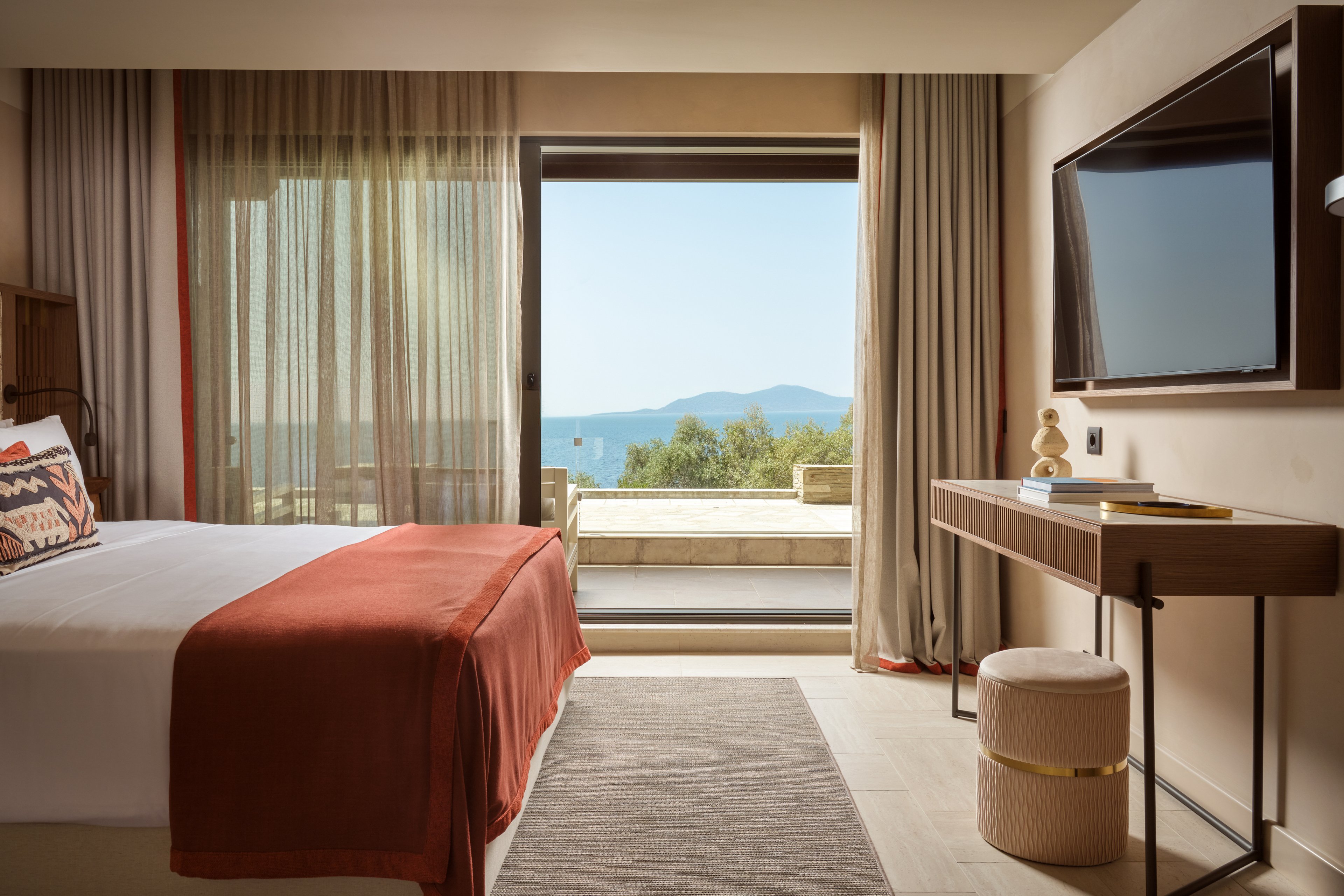 A contemporary hotel room featuring a neatly made bed with a red throw blanket and patterned pillow. The room includes a wall-mounted TV, a wooden desk, and a stool, with large glass doors opening to a balcony overlooking the ocean and distant mountains. Neutral tones and soft lighting create a calm and inviting atmosphere.