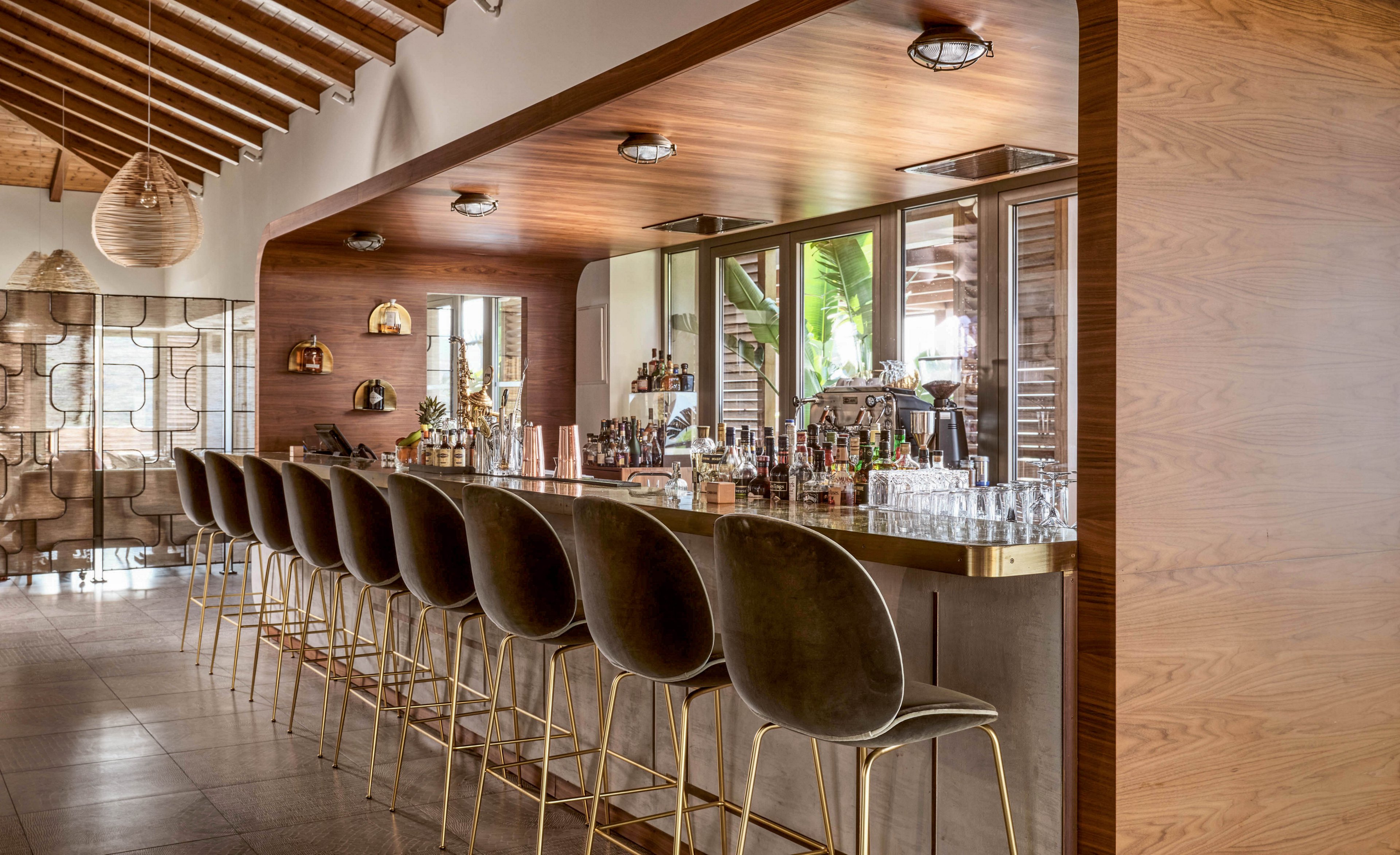 A contemporary bar interior features a long wooden counter with a polished metallic edge and a row of elegant bar stools. The space is empty of patrons, highlighting the clean lines of the furniture and the neatly arranged bottles and glassware behind the bar. Warm wood tones, soft lighting, and exposed ceiling beams create a cozy yet sophisticated atmosphere. Large windows and decorative partitions add depth and a sense of openness to the scene.
