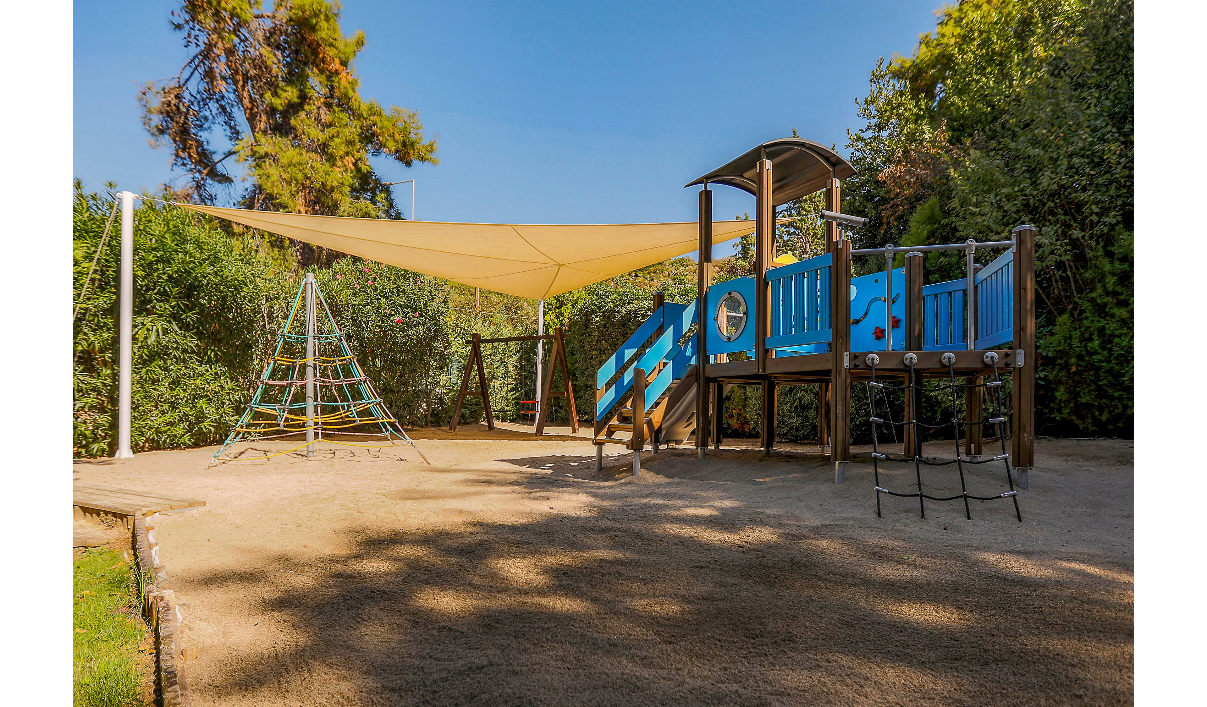 An empty outdoor playground features a blue wooden play structure with slides and platforms. A rope climbing pyramid stands nearby on a sandy ground surface. Large shade sails stretch above the play area, casting soft shadows. Lush green trees and bushes surround the playground, creating a calm and inviting environment under clear daylight.