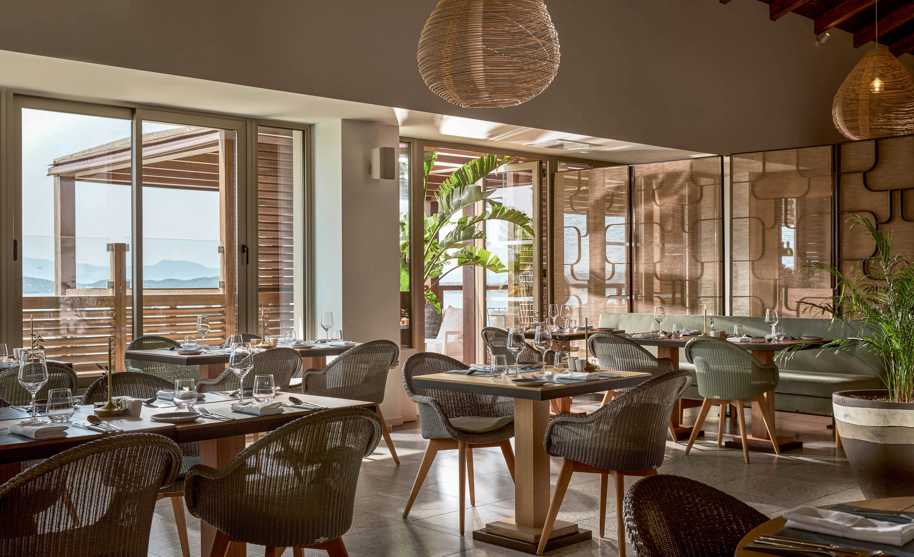 A contemporary restaurant dining room is set with neatly arranged tables and chairs. Large sliding glass doors open to a terrace with wooden structures and a view of the sea in the distance. Soft natural light fills the space, highlighting woven pendant lights, green upholstered seating, and potted plants. The overall atmosphere feels relaxed, airy, and inviting for guests.