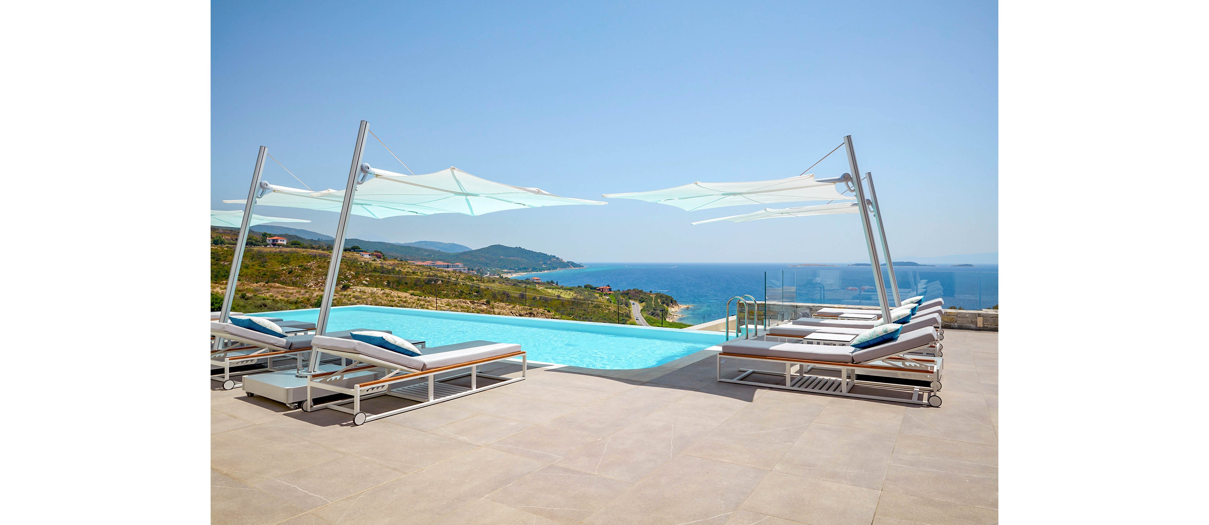 A modern infinity pool sits on a spacious terrace overlooking a scenic coastline and blue sea. Sleek sun loungers with cushions line both sides of the pool under large white shade canopies. The clear sky and calm water create a serene, luxurious resort atmosphere. Rolling hills and distant buildings are visible in the background, emphasizing the elevated vantage point. The overall scene suggests relaxation, leisure, and upscale outdoor living.