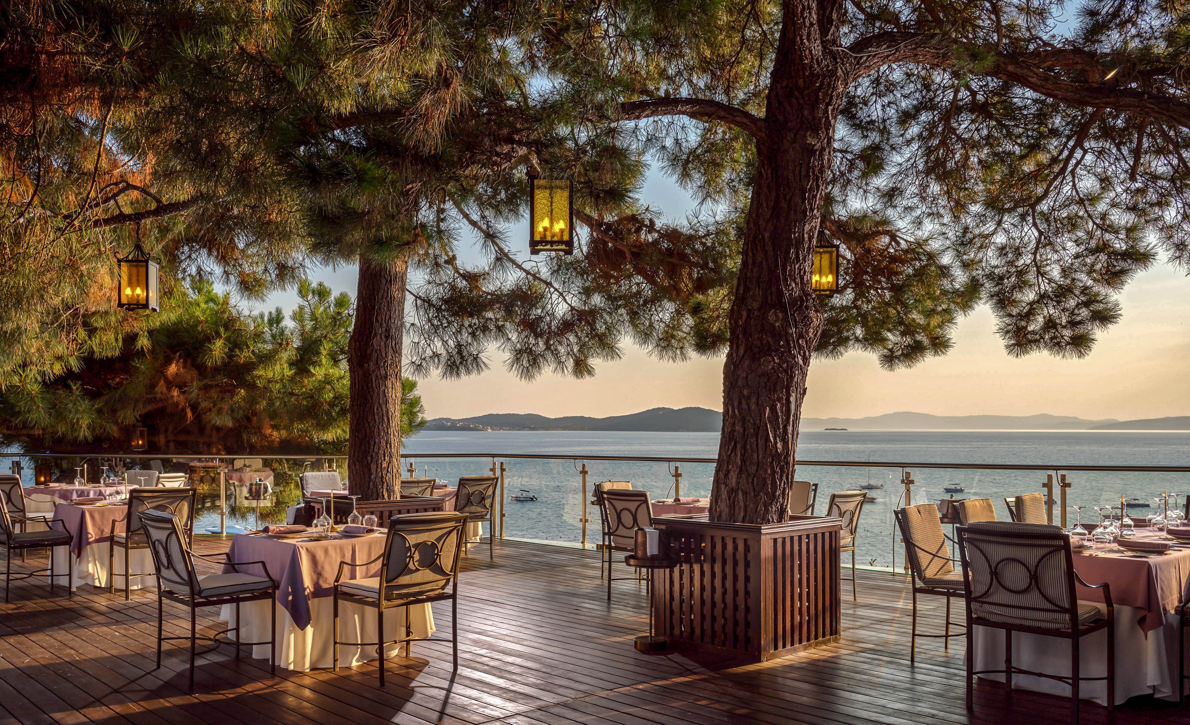 An elegant open-air restaurant terrace overlooks a calm sea at sunset. White tablecloths and neatly arranged place settings sit on a wooden deck beneath tall pine trees. Warm lanterns hang from the branches, casting a soft glow over the dining area. The tranquil water and distant shoreline create a romantic, relaxed atmosphere for outdoor dining.