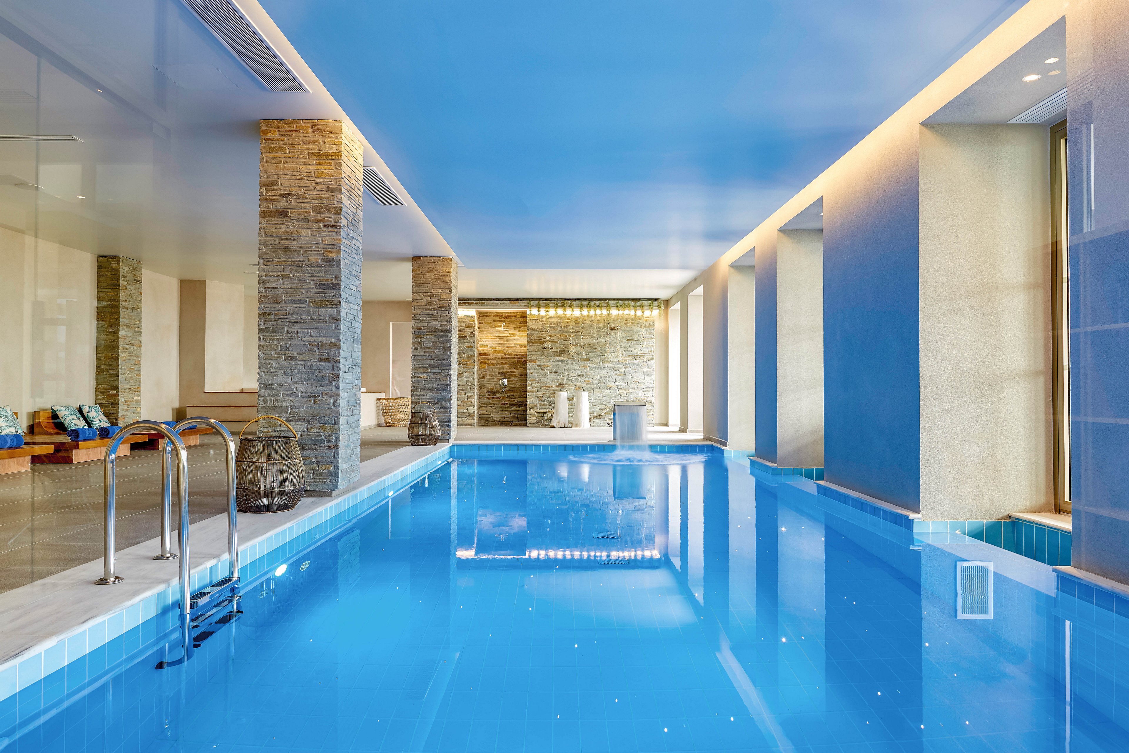 A contemporary indoor swimming pool stretches along a bright, minimalist room. Stone columns and large windows line the pool, allowing natural light to reflect off the calm blue water. Stainless steel ladders and clean tiled edges emphasize a sleek, modern design. Soft seating and warm accent lighting create a relaxing spa-like atmosphere.