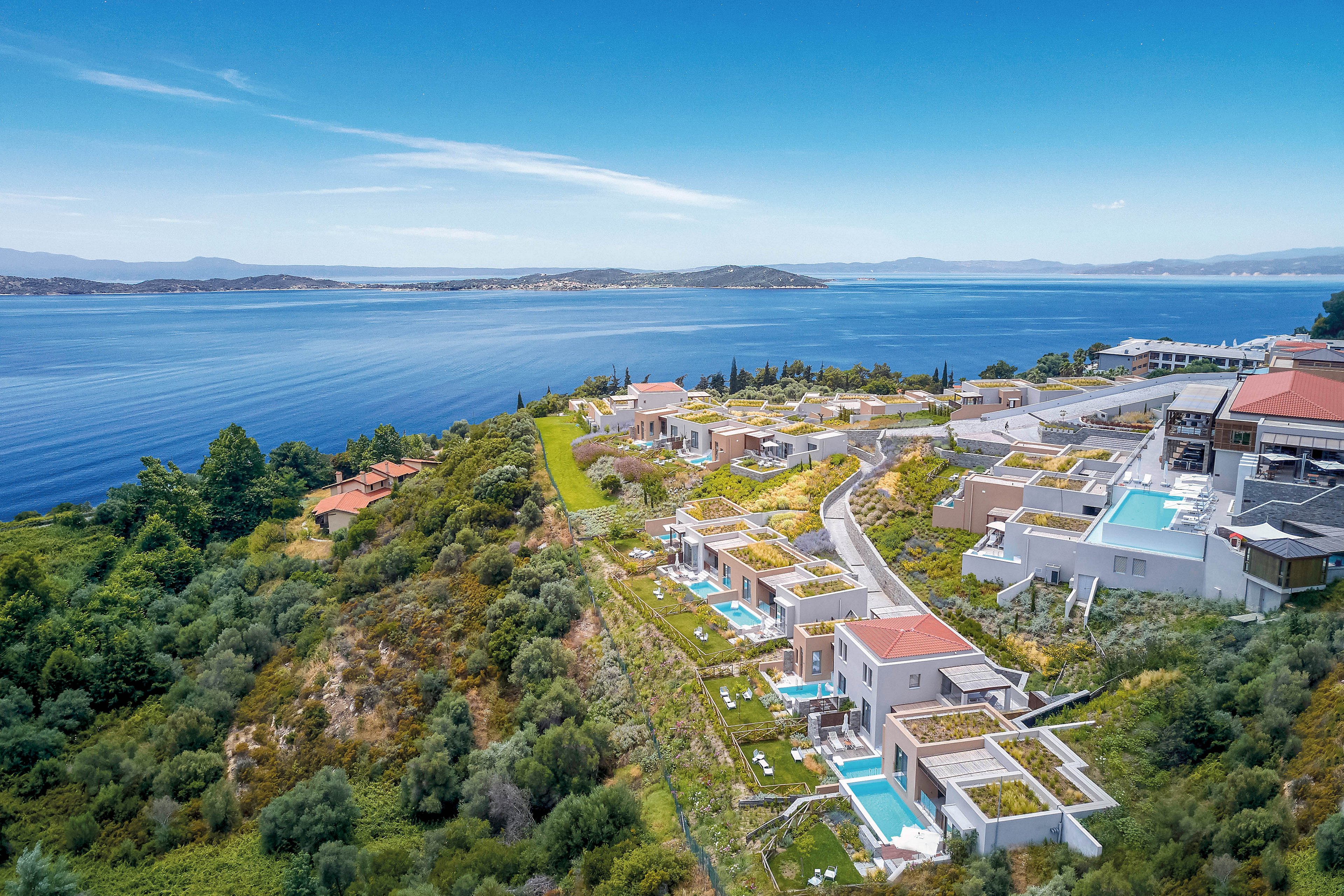 A modern resort complex is built along a lush green hillside overlooking a calm blue sea. Multiple villas with flat roofs and private swimming pools are arranged in tiers beside a winding road. The scene captures a sunny day with clear skies, distant mountains, and scattered houses along the coastline. The contrast between the turquoise pools, greenery, and deep blue water creates a serene vacation atmosphere.