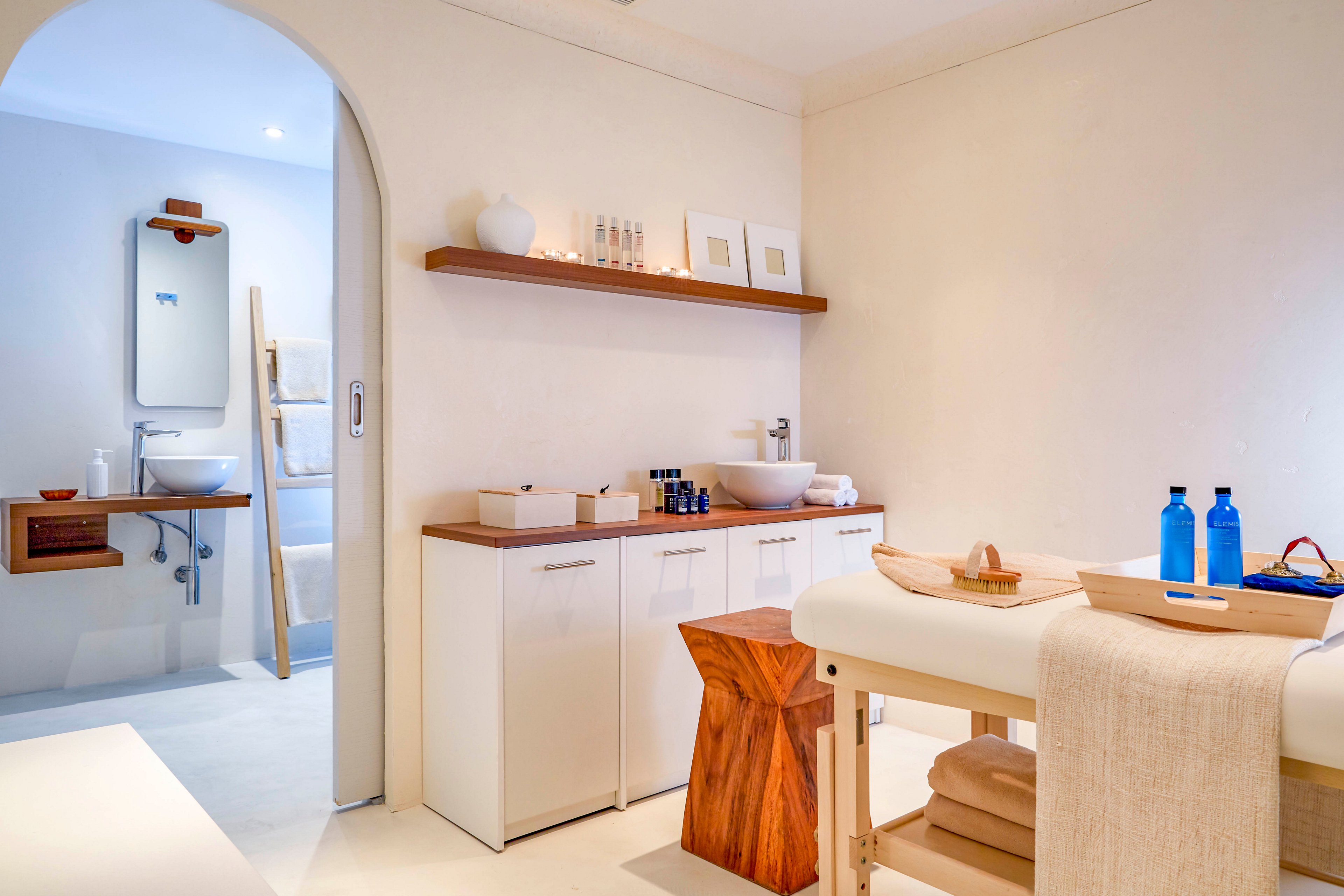 A serene spa treatment room features a massage table covered with neutral linens and folded towels. The space is decorated in a minimalist style with white walls, wooden accents, and a simple wooden stool. Blue bottles and small toiletry containers are neatly arranged on a countertop and tray, adding subtle color. In the background, an open doorway reveals a clean bathroom area with a sink and mirror, enhancing the calm, wellness-focused atmosphere.