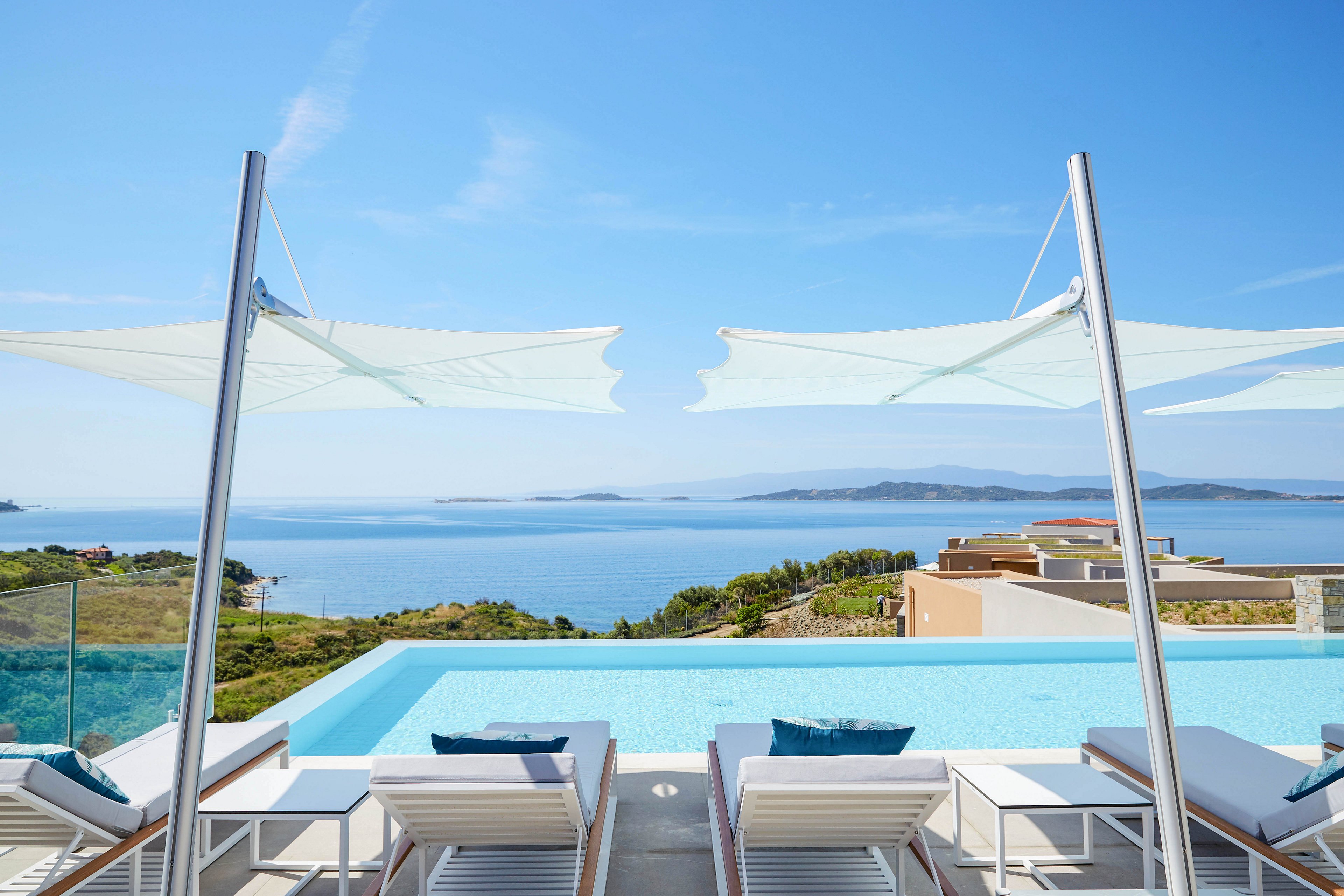 A modern infinity pool terrace overlooks a calm blue sea and distant islands. White lounge chairs with cushions are arranged neatly along the pool edge under sleek fabric shade canopies. Contemporary glass railings frame the view, revealing coastal hills and scattered villas below. The clear sky and bright sunlight create a serene, luxurious vacation atmosphere.
