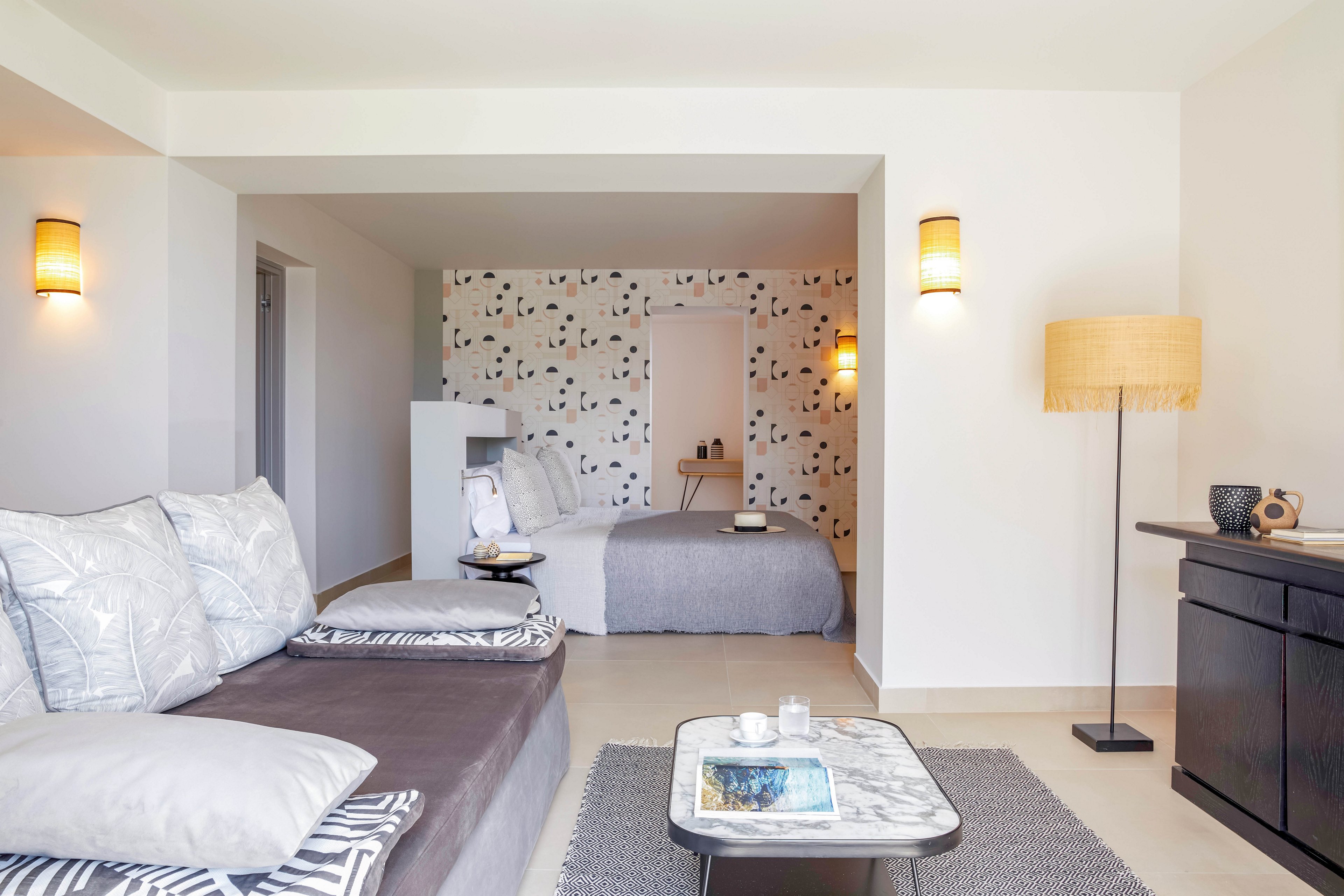 A contemporary hotel suite features a cozy lounge area with a cushioned daybed and patterned pillows in the foreground. In the background, a neatly made bed with neutral bedding sits against a decorative accent wall with abstract shapes. Warm wall sconces and a floor lamp provide soft ambient lighting that complements the light-colored walls and flooring. A small coffee table with books and candles and a dark sideboard with decorative objects complete the minimalist, modern decor.