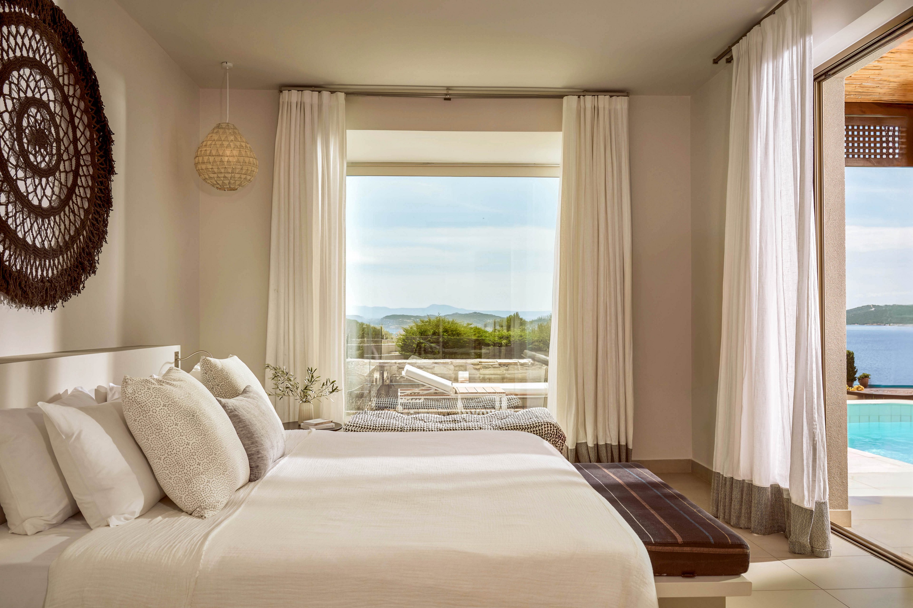 A modern bedroom features a neatly made bed with white linens and plush pillows. Large floor-to-ceiling curtains frame a wide window that looks out onto a terrace and distant sea. Natural light fills the room, highlighting neutral tones and minimal decor, including a woven wall hanging and small bedside plants. An open doorway reveals a pool area and blue water, enhancing the serene, vacation-like atmosphere.