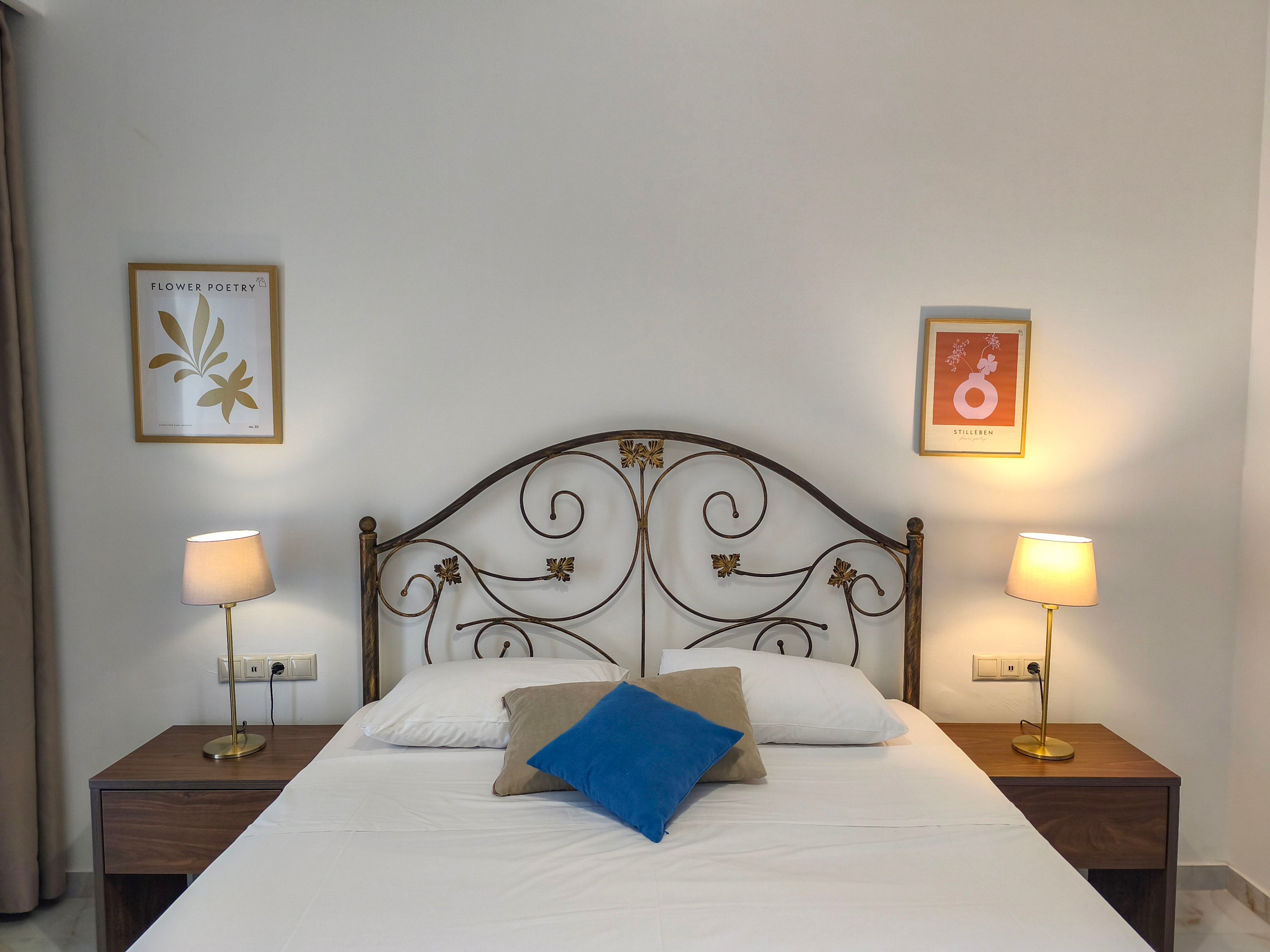 A neatly made double bed with white linens is centered against a plain light-colored wall. The bed features an ornate metal headboard with swirling patterns and small decorative accents. Two wooden bedside tables with matching gold lamps flank the bed, casting a warm glow. Framed artwork hangs above each nightstand, and a trio of accent pillows, including a blue one, adds a touch of color to the calm, minimalist space.