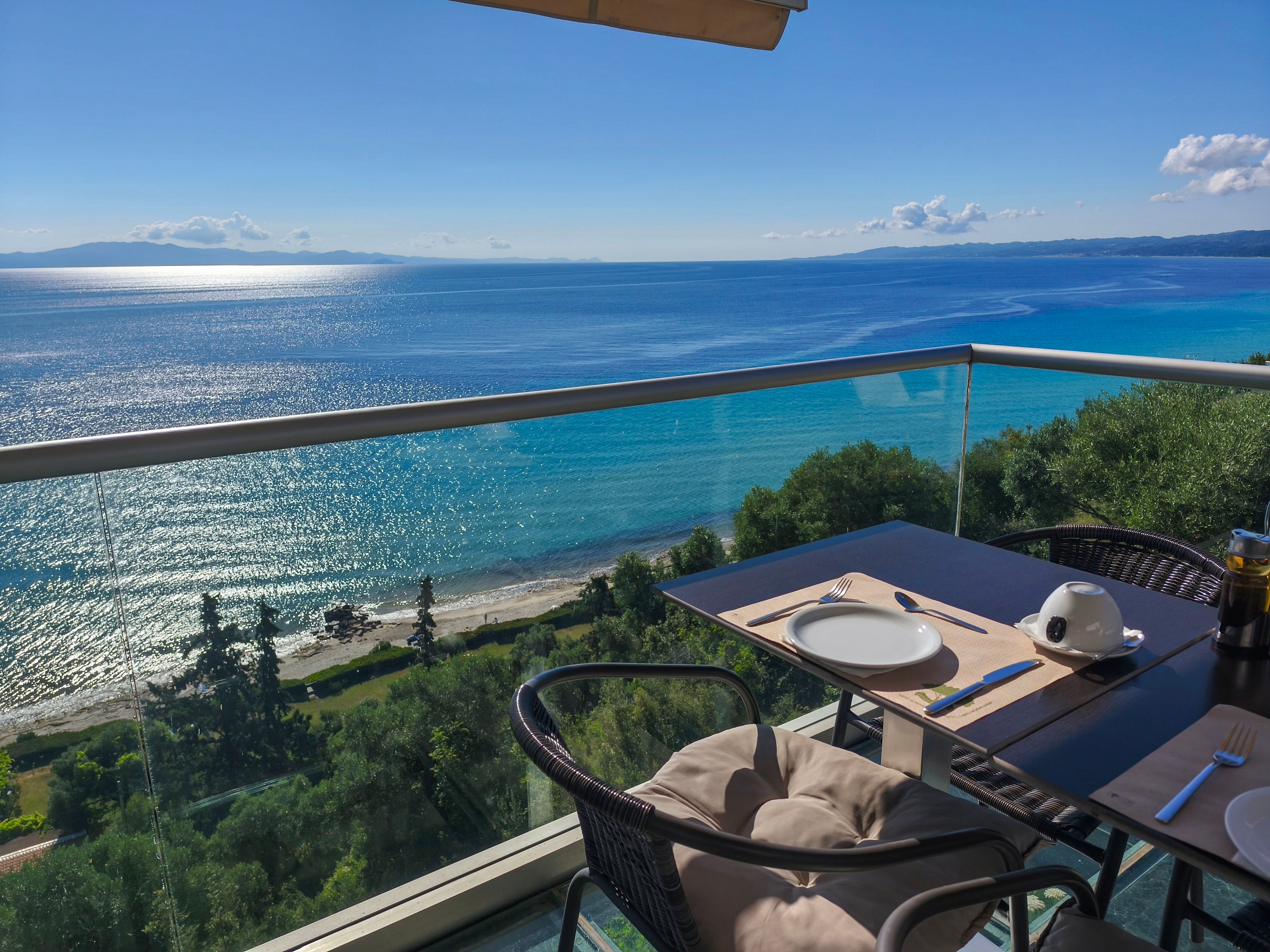 A restaurant terrace table is set for two overlooking a stunning coastal landscape. The scene features clear blue ocean water, gentle waves, and a distant horizon under a bright, sunny sky. Green trees and a sandy shoreline stretch below the glass balcony, adding depth and contrast to the view. The overall atmosphere feels tranquil and inviting, ideal for a relaxed seaside meal.