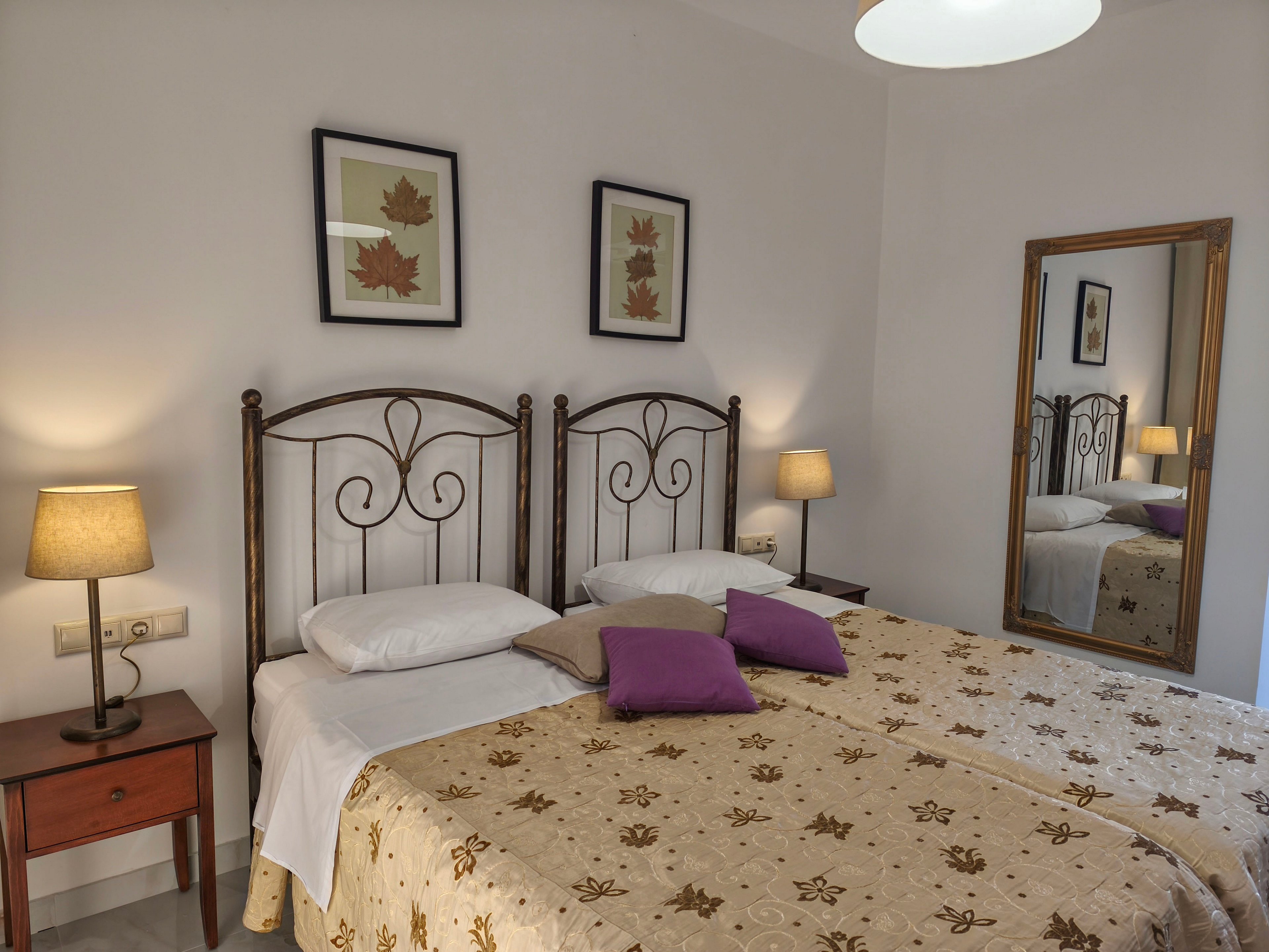 A cozy bedroom features two twin beds pushed together with ornate metal headboards. The beds are covered with beige patterned bedspreads and accented by purple throw pillows. Warm table lamps on wooden nightstands illuminate the room, creating a soft, inviting atmosphere. Wall art with leaf motifs and a large framed mirror add decorative touches to the simple, modern interior.