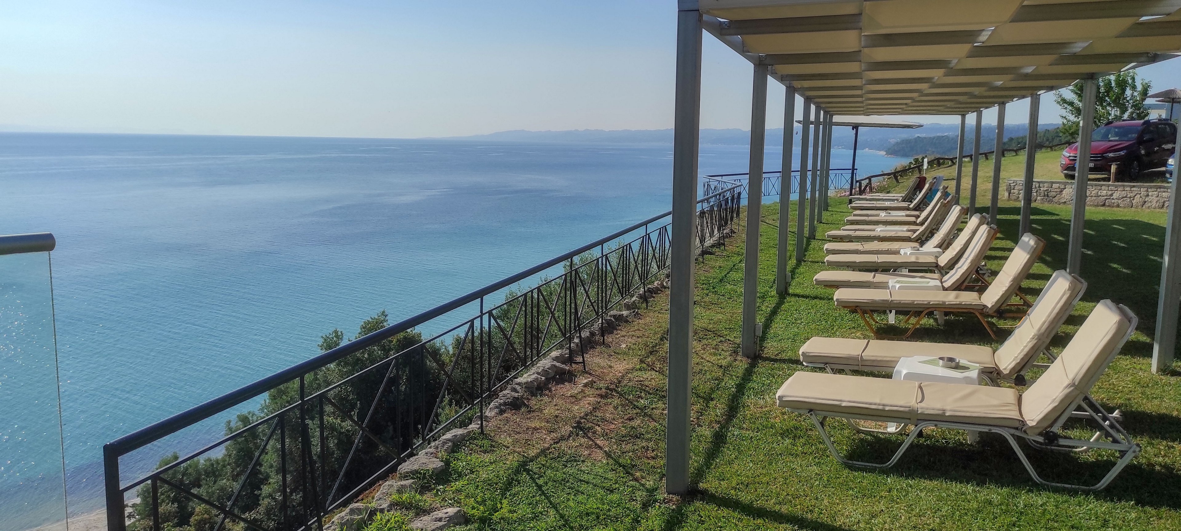 A row of empty lounge chairs is arranged under a pergola on a grassy terrace overlooking a calm, expansive sea. The metal railing runs along the edge of the cliff, providing a clear view of the coastline and distant horizon. Bright sunlight casts shadows from the pergola structure, emphasizing the orderly alignment of the chairs. The scene suggests a peaceful resort or vacation setting with a relaxed, tranquil atmosphere.