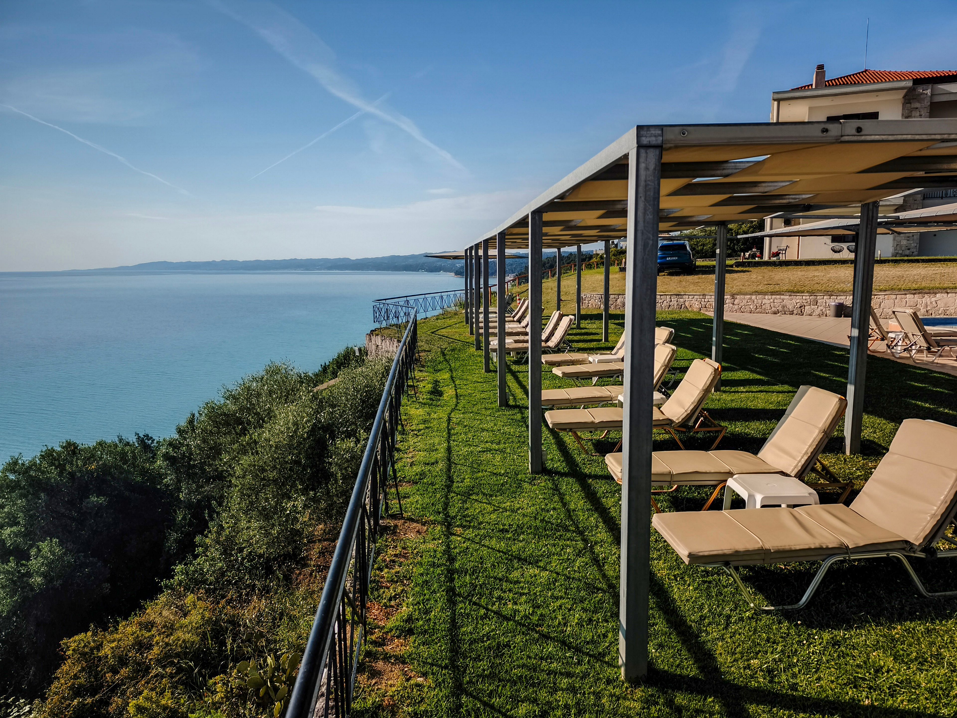 A row of beige lounge chairs sits neatly under a metal pergola on a grassy terrace overlooking the sea. The terrace is bordered by a black railing and drops down to lush green vegetation on the cliffside. Calm blue water stretches to the horizon under a clear sky with faint contrails. A nearby building and additional seating area suggest a coastal resort or hotel setting. The scene feels quiet and inviting, ideal for sunbathing and relaxation.