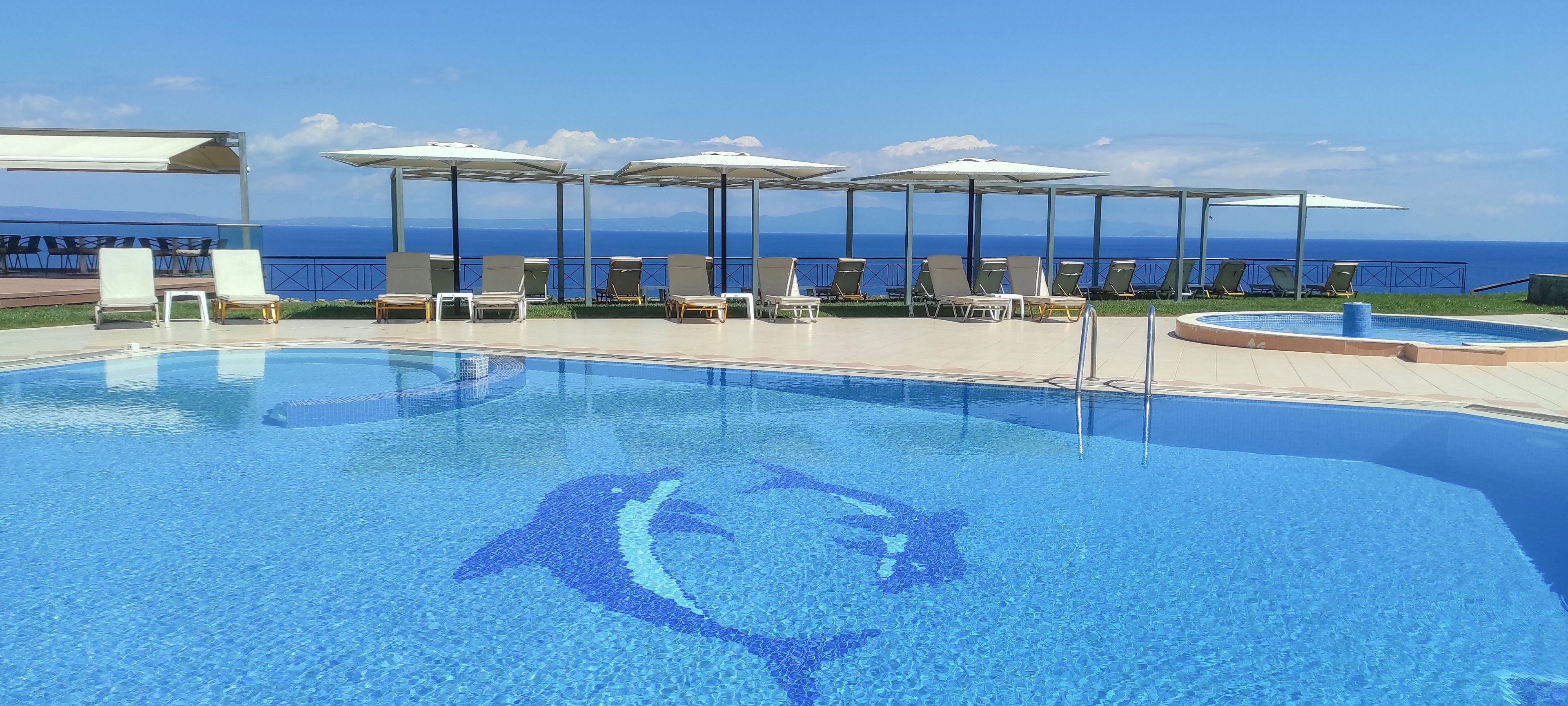 An outdoor swimming pool with a blue dolphin mosaic dominates the foreground of this seaside resort scene. Sun loungers and white parasols line the poolside terrace, facing the calm blue ocean in the background. The sky is clear with a few scattered clouds, enhancing the tranquil coastal atmosphere. Metal handrails and a clean tiled deck emphasize the modern, well-maintained design of the pool area.