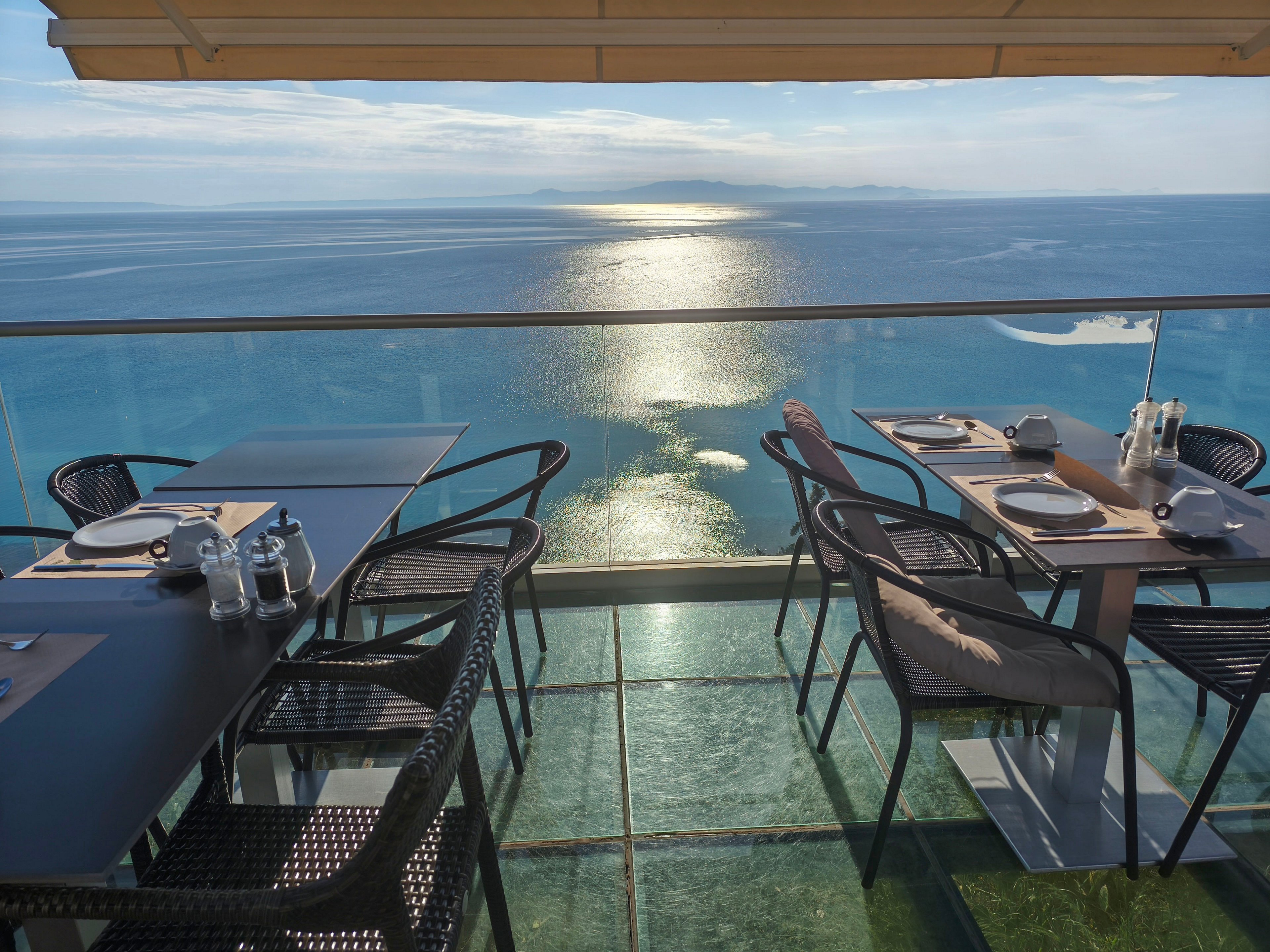 An elegant rooftop restaurant terrace overlooks a calm, expansive ocean at sunset. Empty tables are neatly set with plates, cutlery, glasses, and condiments, ready for guests. The glass floor and clear railing enhance the dramatic view of the shimmering water and distant horizon. Soft natural light and a large overhead awning create a relaxed, upscale dining atmosphere.