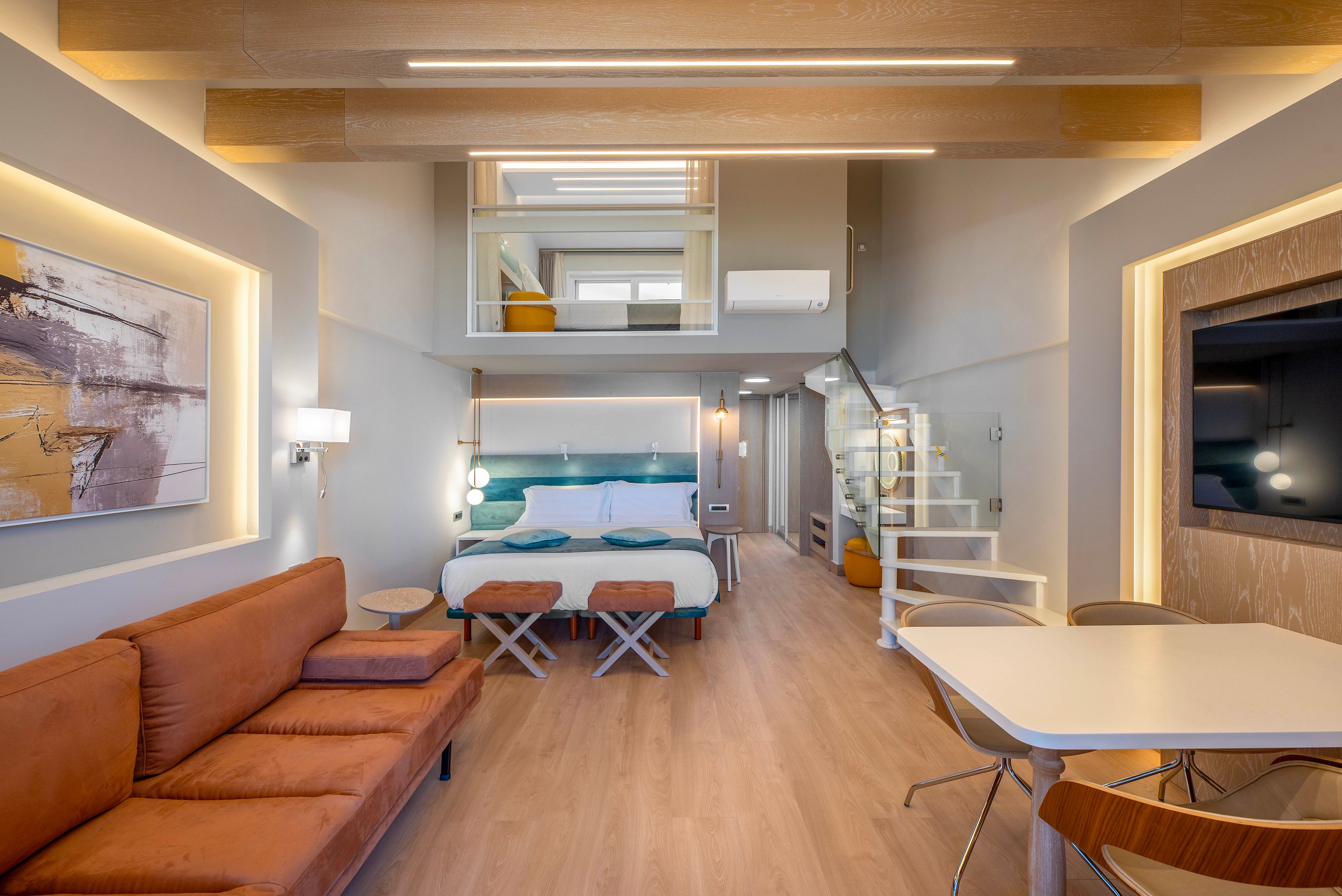 Contemporary loft-style hotel room featuring a central double bed with teal accents and two small benches at the foot. A brown leather sofa lines one wall opposite a mounted flat-screen television and a white dining table with wooden chairs. A spiral staircase leads up to a mezzanine level, adding vertical interest and extra space. Warm wood tones, recessed lighting, and abstract wall art create a bright, inviting atmosphere.