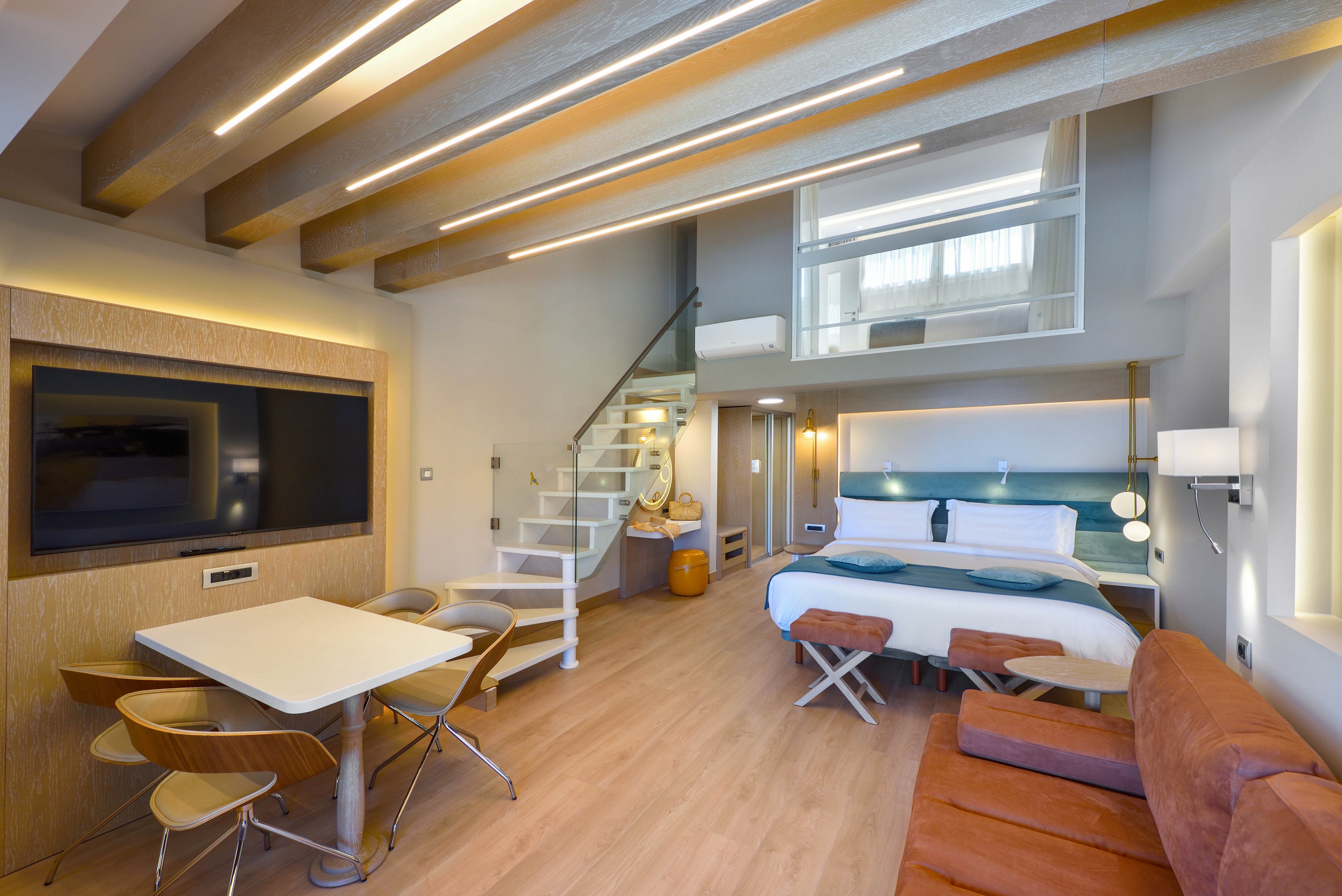 Contemporary loft-style hotel room featuring a double bed and a cozy sitting area. A small dining table with chairs sits near a large flat-screen television mounted on the wall. A white spiral staircase leads up to a mezzanine level, adding vertical interest to the space. Warm wood flooring, soft ambient lighting, and clean minimalist decor create a bright and inviting atmosphere.