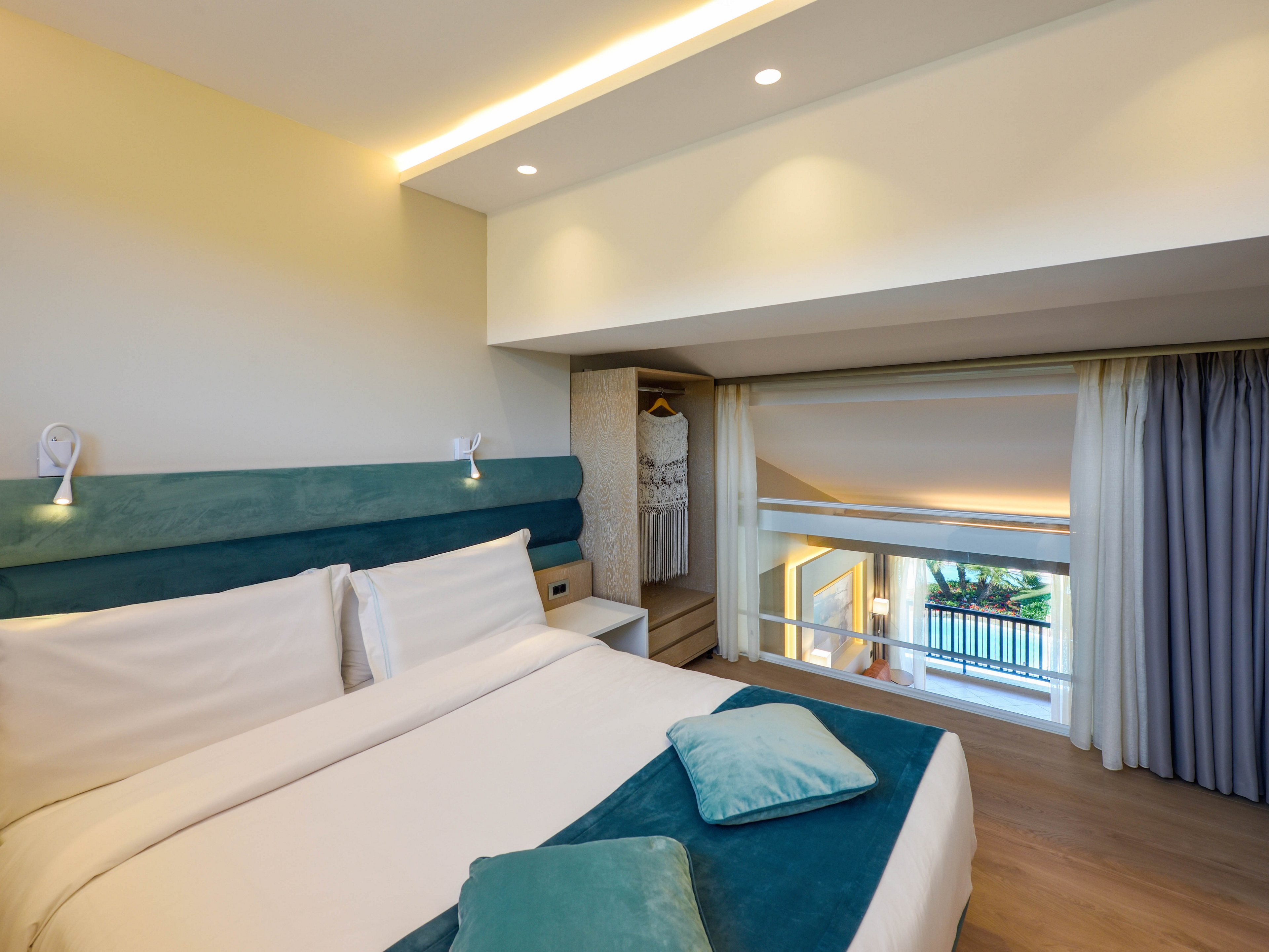 Contemporary hotel bedroom featuring a large neatly made bed with teal accents and decorative pillows. The room opens to a loft-style interior balcony overlooking a bright lower level with large windows. Soft recessed lighting and neutral walls create a calm, inviting atmosphere. Sheer and blackout curtains frame the view, while a small open wardrobe adds functional detail.