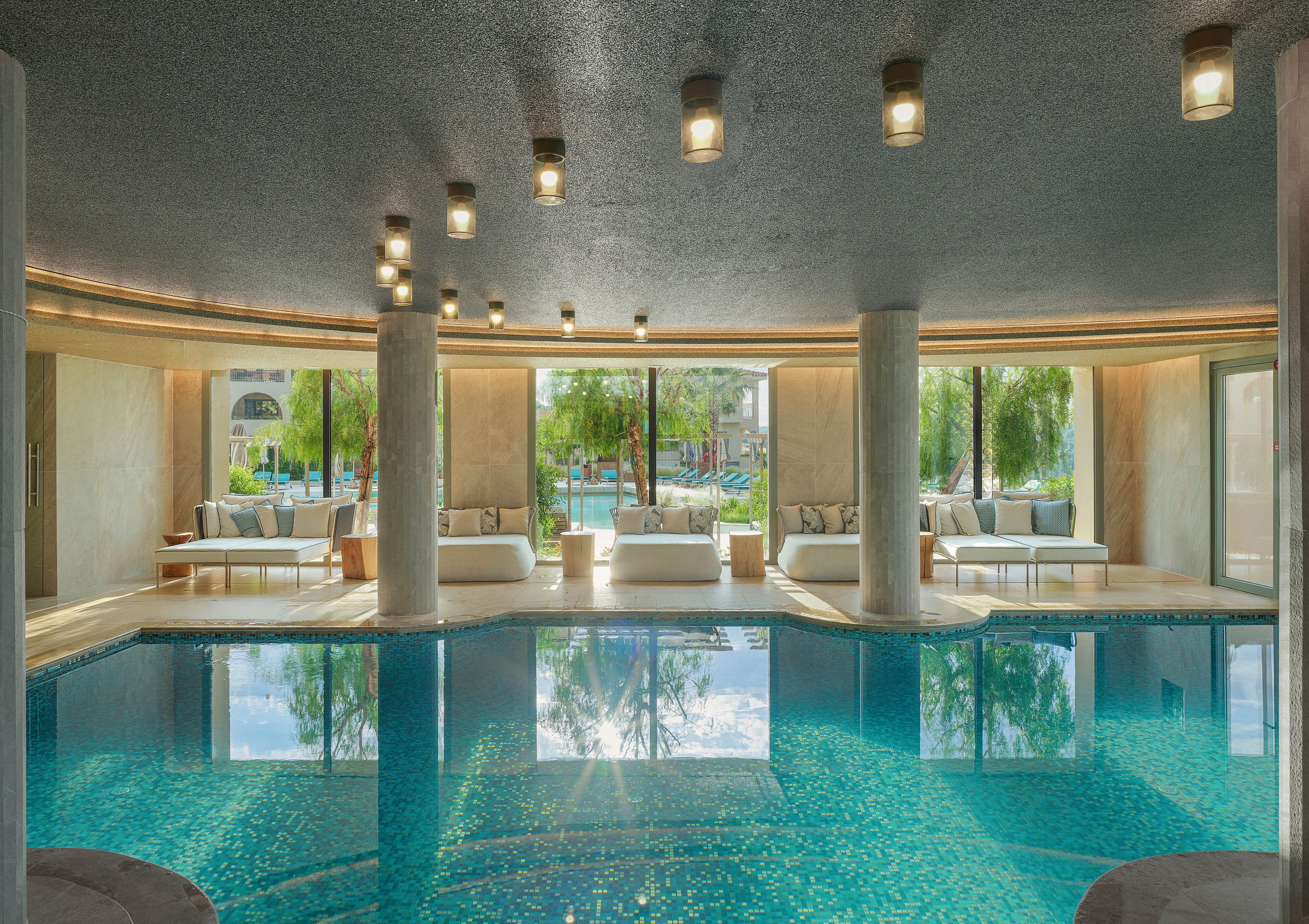 A serene indoor swimming pool is surrounded by modern columns and soft seating areas. Large floor-to-ceiling windows reveal lush greenery and an outdoor pool beyond. Warm ceiling lights reflect on the calm turquoise water, creating a tranquil atmosphere. Neutral-toned walls and cushioned daybeds enhance the spa-like, contemporary design.