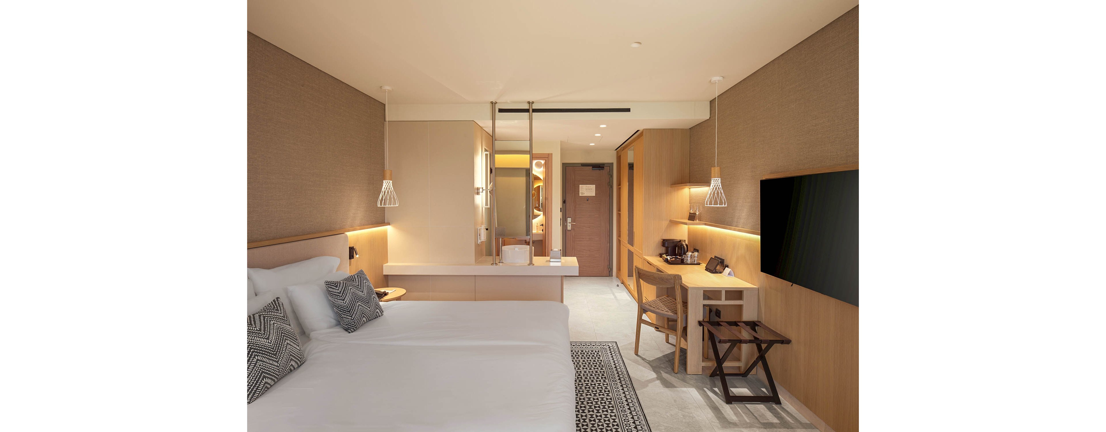 A contemporary hotel room features a large neatly made bed with white linens and patterned pillows. The space includes a wooden desk with a chair, a flat-screen television mounted on the wall, and a small hospitality station with a kettle and cups. Warm recessed lighting and pendant lamps create a cozy atmosphere, highlighting the neutral color palette and clean lines. A hallway leads to the entrance door and bathroom area, giving the room a spacious, organized feel.