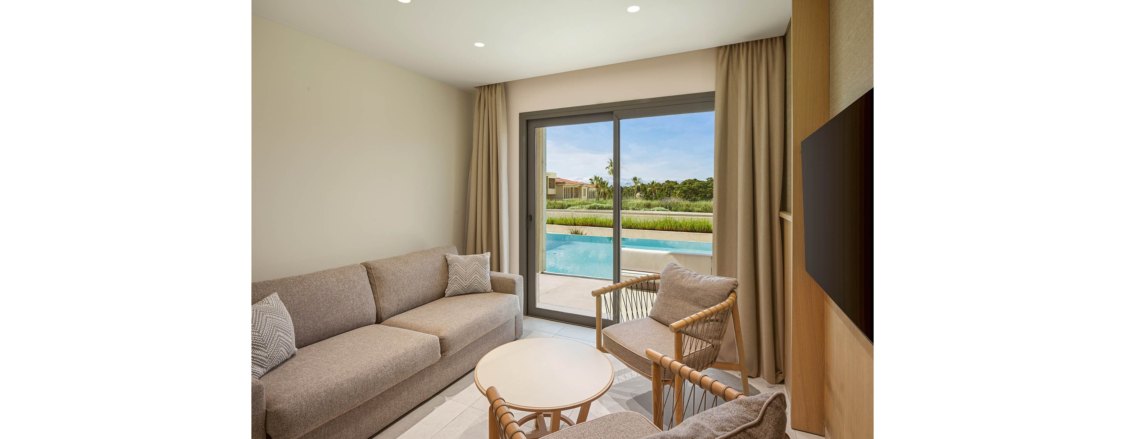 A contemporary living room features a beige sofa, armchairs, and a small round coffee table. Large sliding glass doors open to a private swimming pool and landscaped garden area. Neutral tones and soft lighting create a calm, minimalist atmosphere. The scene suggests a comfortable vacation suite or resort accommodation with an inviting outdoor space.