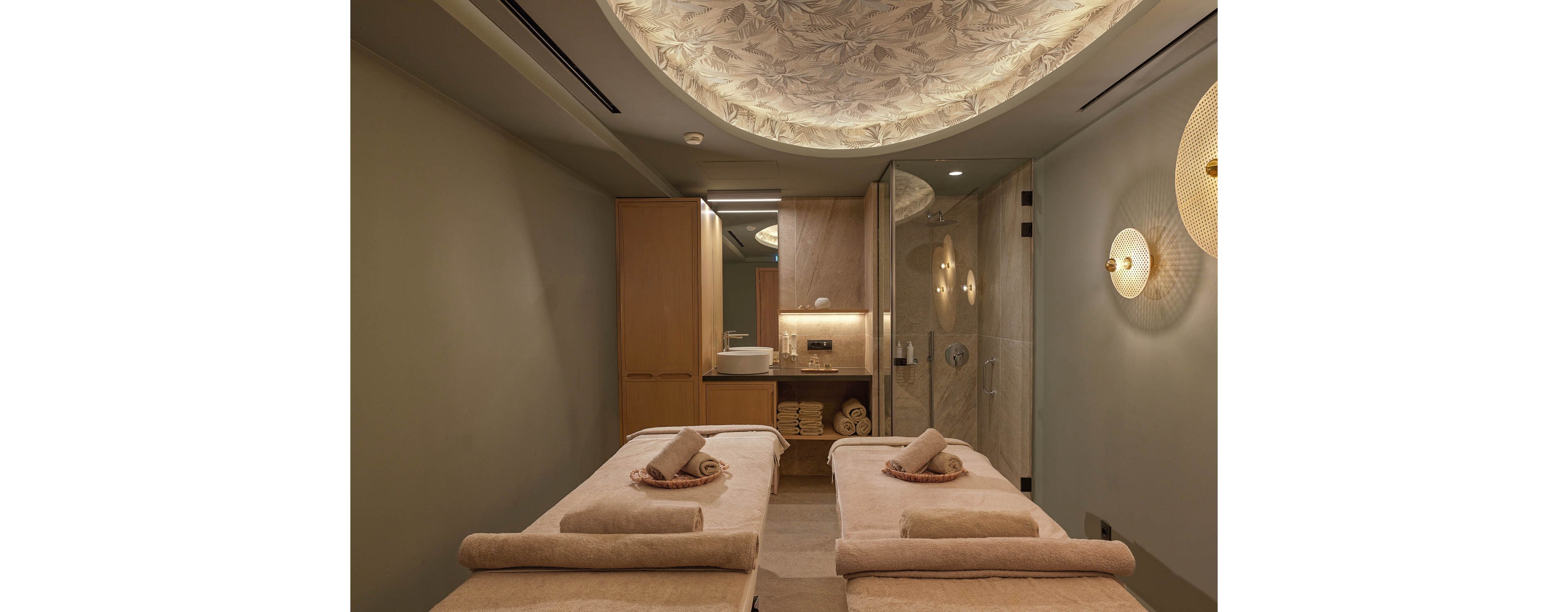 A serene spa treatment room features two massage beds neatly prepared with rolled towels. Soft, warm lighting and a decorative ceiling create a calming atmosphere. Wooden cabinetry and shelves with stacked towels add to the organized, minimalist design. A glass-enclosed shower area is visible at the back, enhancing the sense of a complete wellness space.