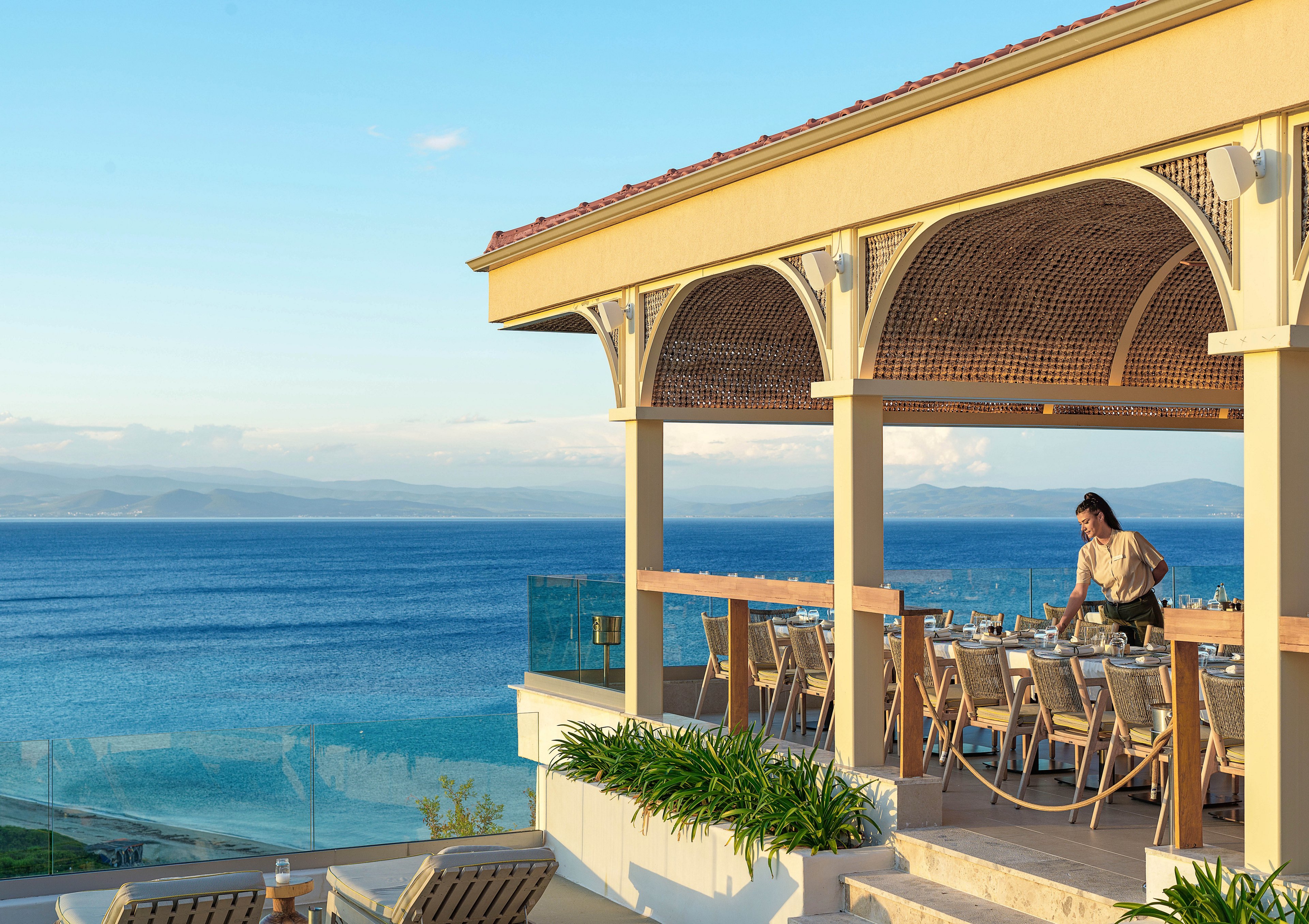 An open-air restaurant terrace sits above a vivid blue ocean, framed by elegant arches and a wooden roof. A staff member arranges tables and chairs, preparing the dining area for guests. The scene is bathed in warm late-afternoon light, with clear skies and distant coastline visible on the horizon. Lush green plants and glass railings add a modern, relaxed resort atmosphere.