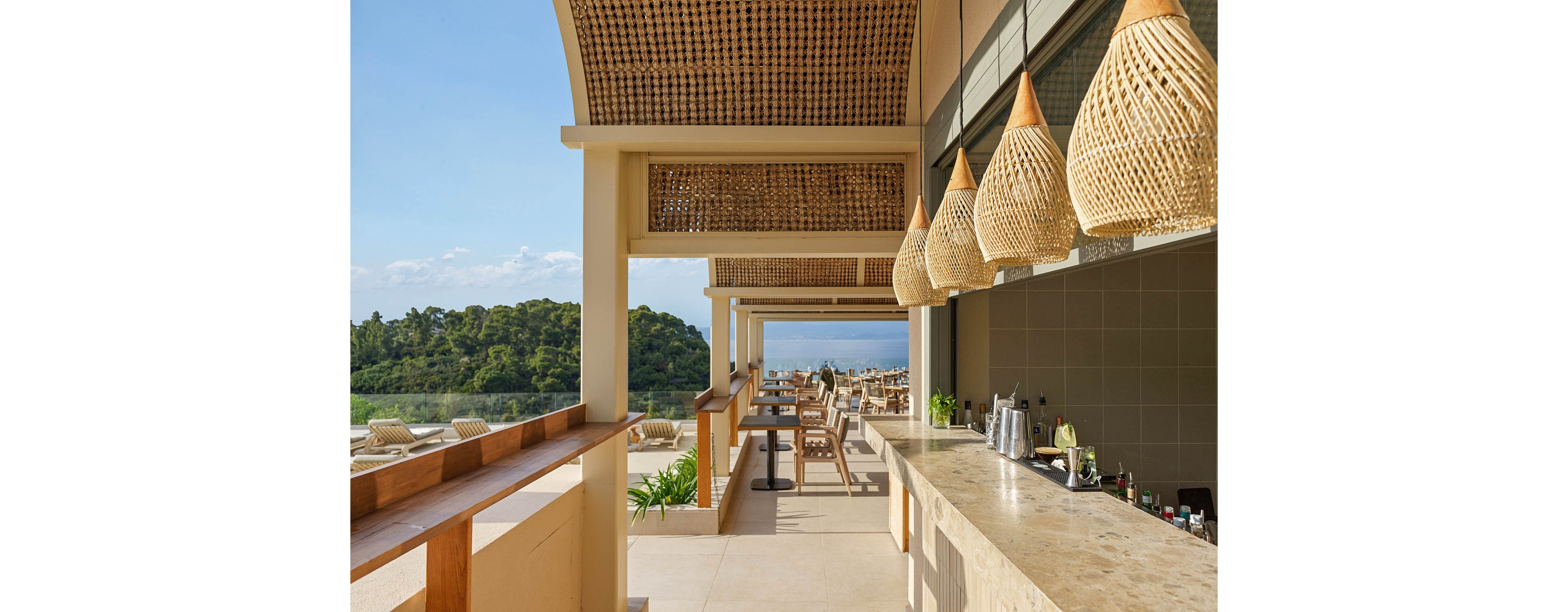 An elegant open-air bar area features a long stone counter with hanging woven pendant lights. Wooden tables and chairs line the terrace, facing toward the distant ocean and clear blue sky. Lush green trees frame the view, enhancing the relaxed coastal atmosphere. The design combines natural materials and neutral tones for a modern, tropical resort feel.