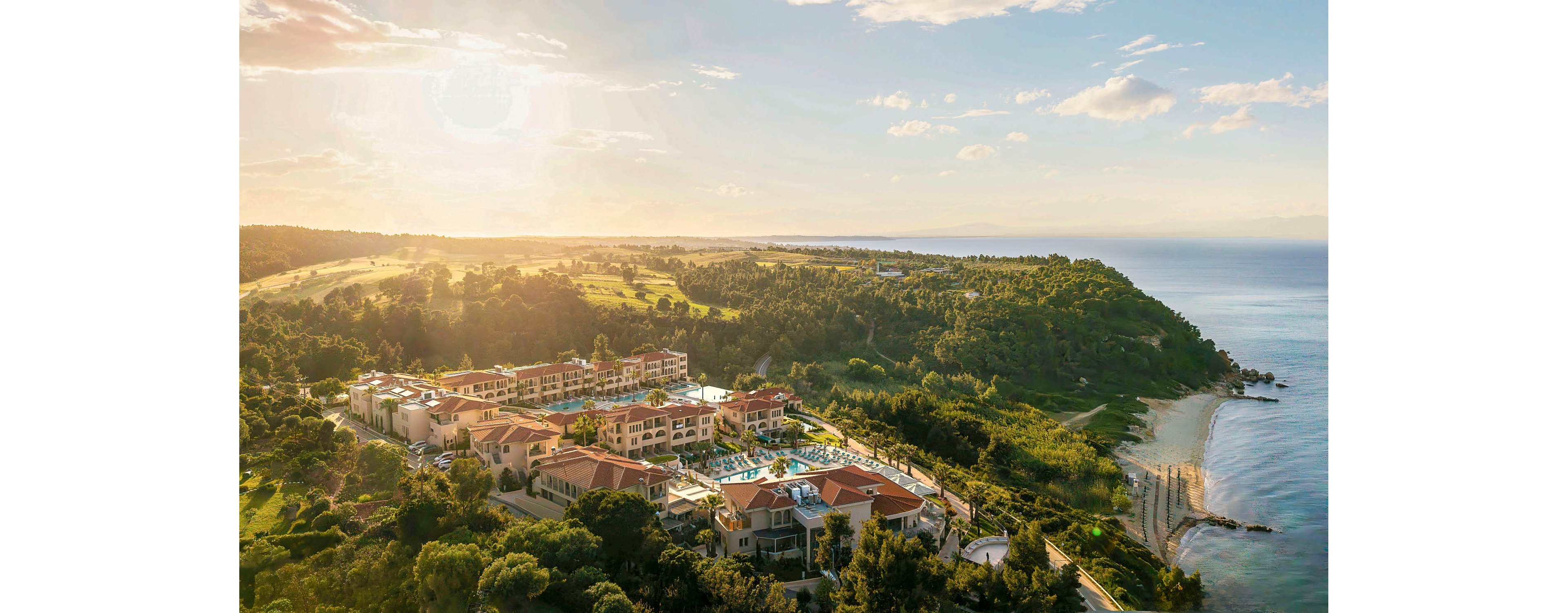A large seaside resort complex sits along a lush green peninsula, bordered by the calm sea and gently sloping hills. Multiple buildings with terracotta roofs surround bright blue swimming pools and landscaped grounds. The coastline curves around sandy beaches and rocky edges, while the sun casts a warm golden light across the scene. The overall atmosphere is tranquil and inviting, suggesting a luxurious vacation destination.