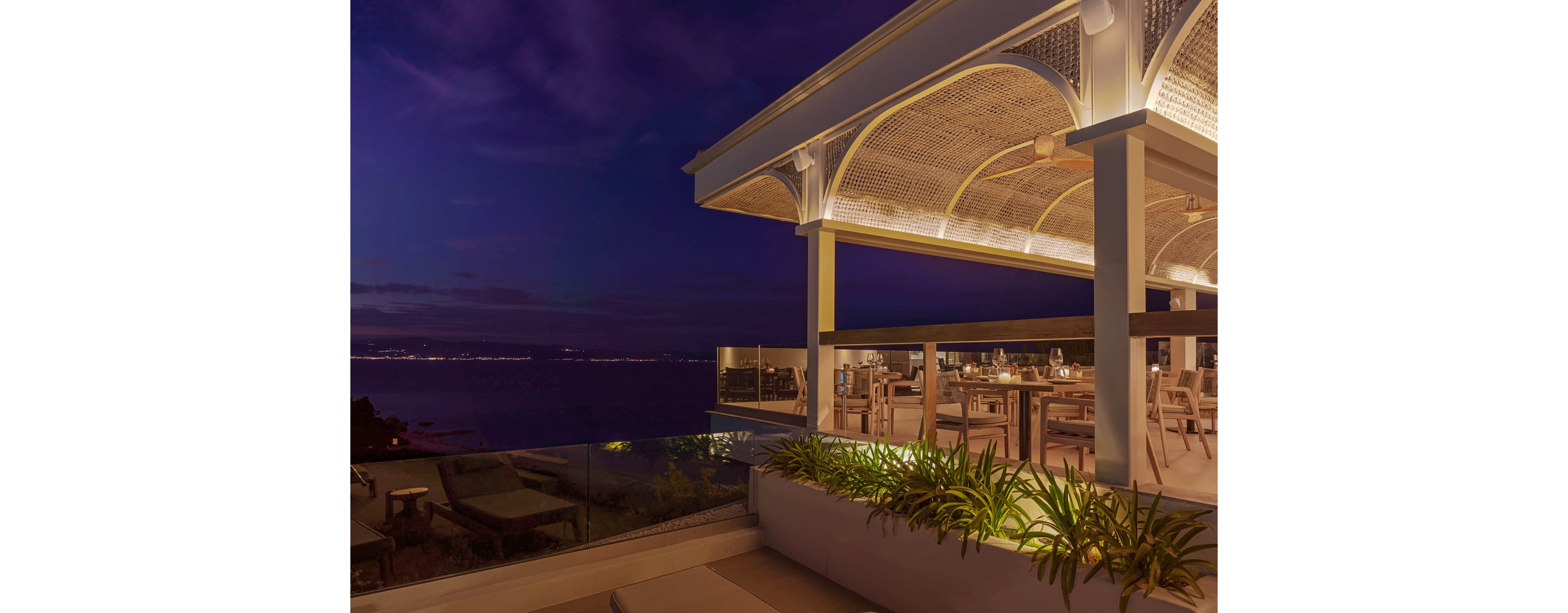 A modern open-air rooftop restaurant overlooks a calm sea at dusk. Warm architectural lighting highlights the arched canopy and neatly arranged dining tables and chairs. Lush green plants line the edge of the terrace beside a glass railing, adding a natural touch to the contemporary design. City lights shimmer faintly along the distant coastline under a deep blue evening sky.