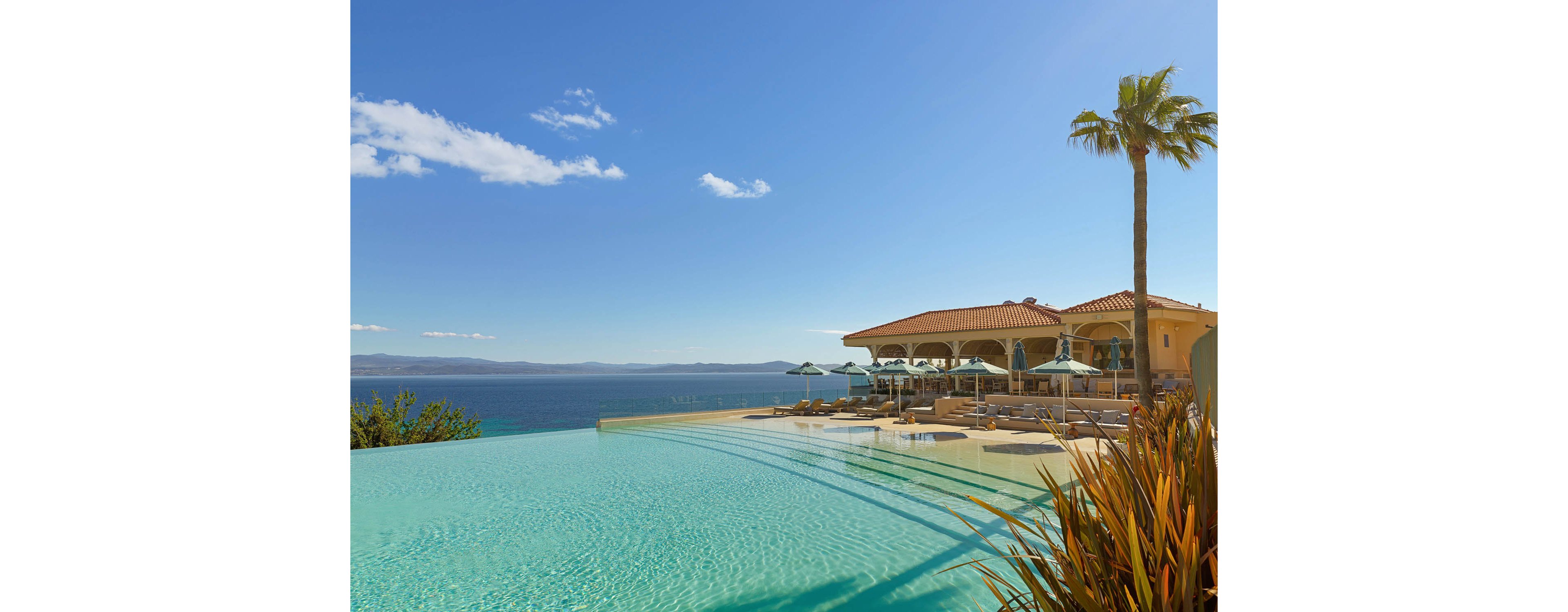 A luxurious infinity pool stretches toward a calm blue ocean under a clear sky. A Mediterranean-style resort building with arches and a terracotta roof sits beside the pool, shaded by umbrellas and lounge chairs. A tall palm tree and ornamental plants frame the scene, adding a tropical touch. The bright sunlight and soft reflections on the water create a serene, vacation-like atmosphere.