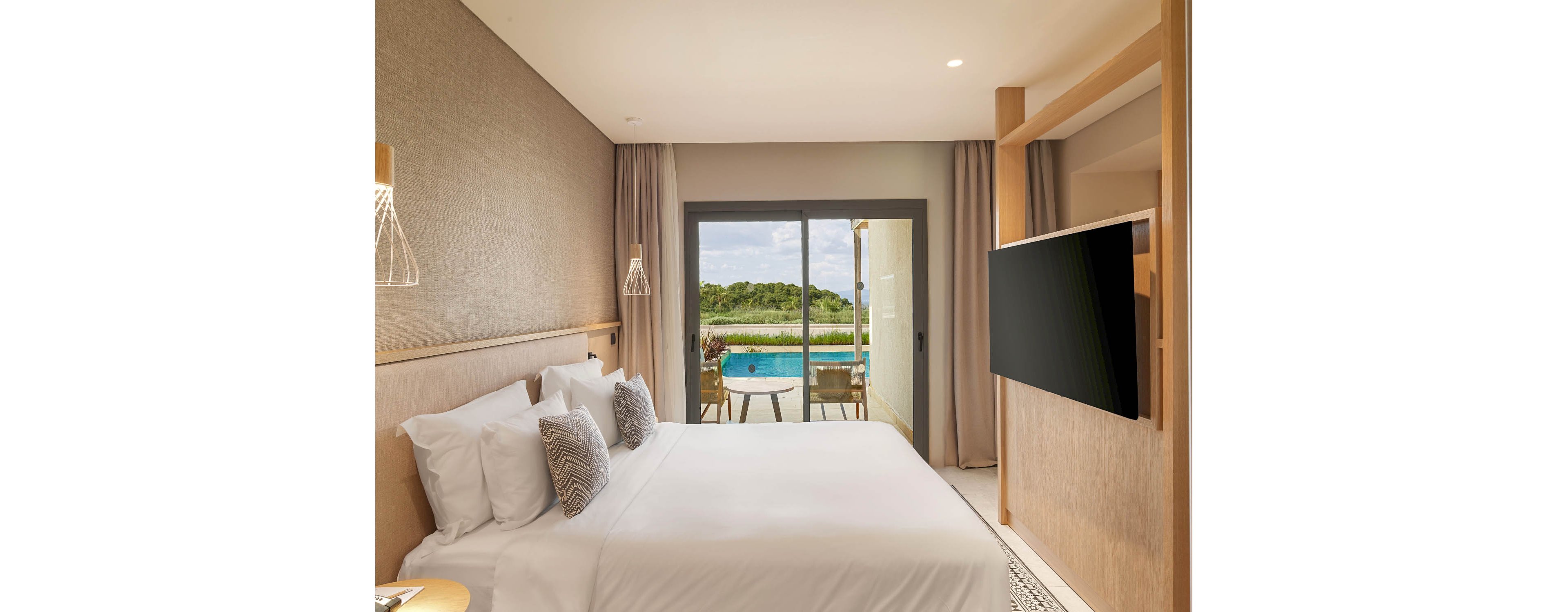 A contemporary hotel bedroom features a neatly made double bed with white linens and accent pillows. Floor-to-ceiling curtains frame a large sliding glass door that opens to a private terrace. Outside, a small plunge pool and patio chairs overlook a lush green landscape under a bright sky. Warm neutral tones, soft lighting, and minimalist decor create a calm, upscale atmosphere.