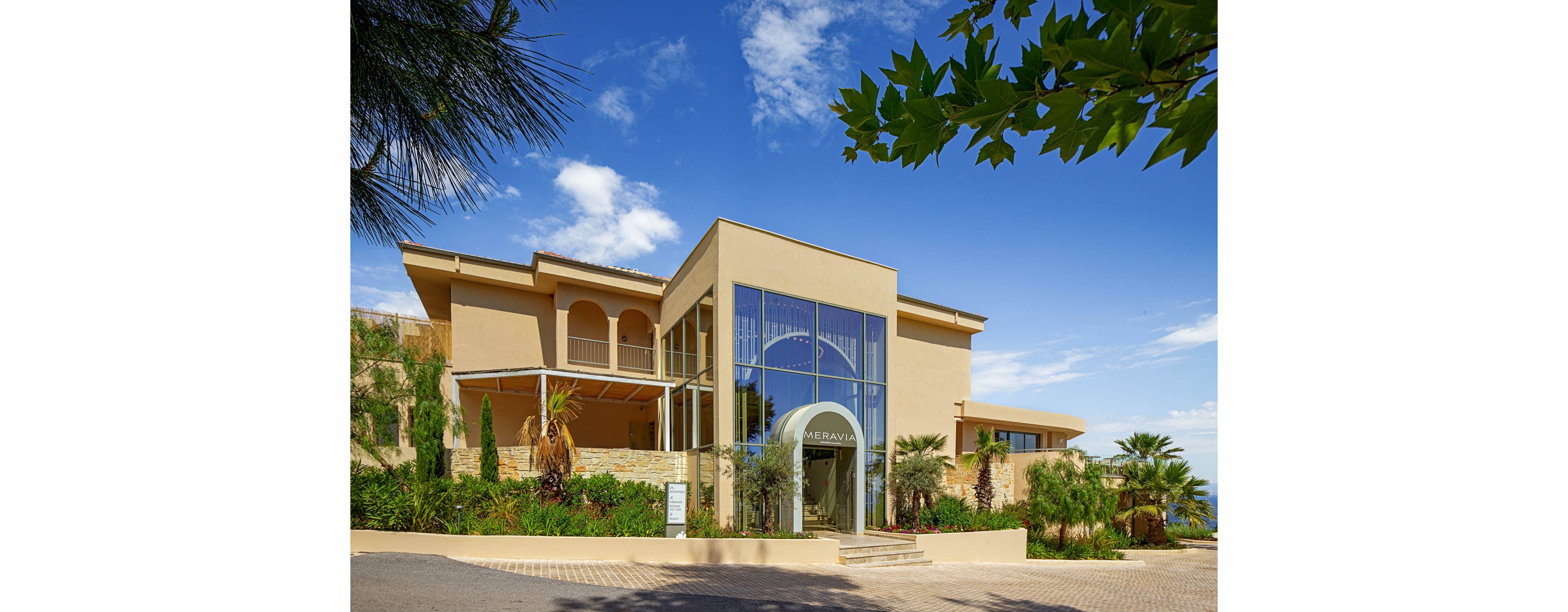 A contemporary hotel building features a prominent glass entrance framed by beige stucco walls. The structure is surrounded by lush landscaping, including palm trees and other greenery, set against a bright blue sky with scattered clouds. Large windows reflect the outdoor scenery, emphasizing the modern architectural style. The paved driveway and clean exterior suggest a welcoming resort or hospitality setting.
