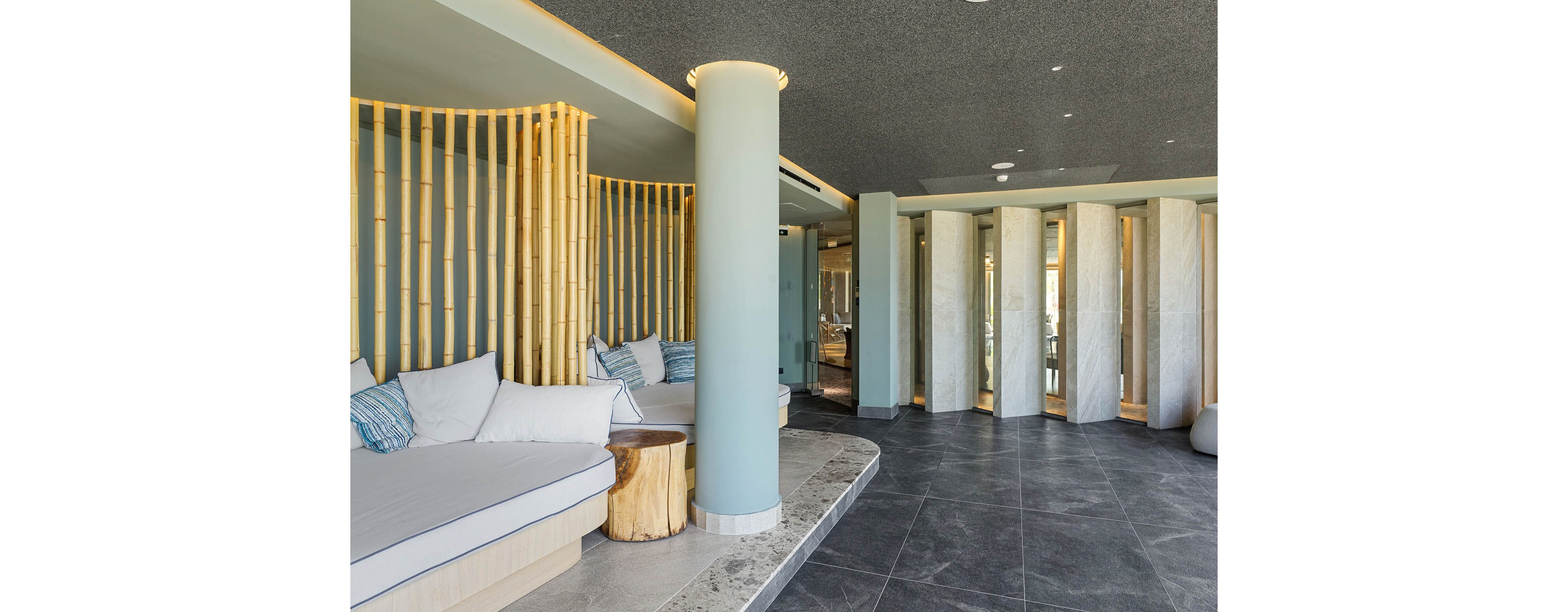 A contemporary spa lounge features built-in cushioned seating with striped and solid pillows. Vertical bamboo slats and rounded columns create a natural, organic partition between areas. The space has dark tiled flooring, soft neutral walls, and warm recessed lighting in the ceiling. Overall, the interior feels calm, minimalist, and inviting for relaxation.