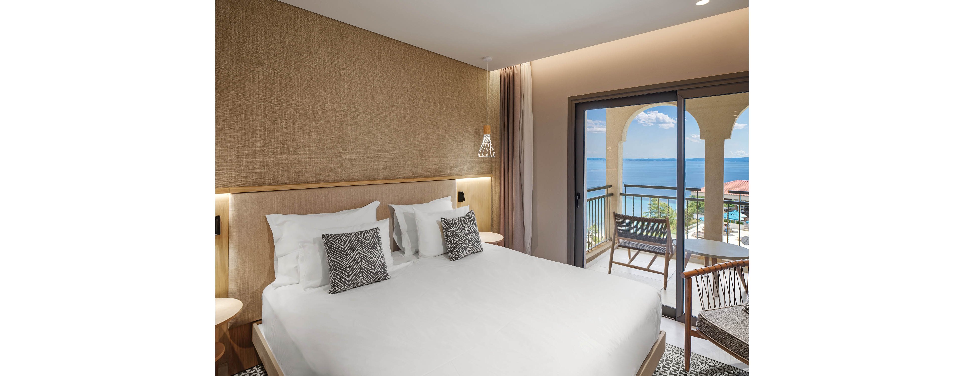 A contemporary hotel bedroom features a large neatly made bed with white linens and patterned accent pillows. Warm neutral walls and soft lighting create a calm, inviting atmosphere. A sliding glass door opens to a balcony with chairs and a table overlooking a bright blue ocean and arched architectural details. The scene suggests a relaxing seaside vacation or resort stay.
