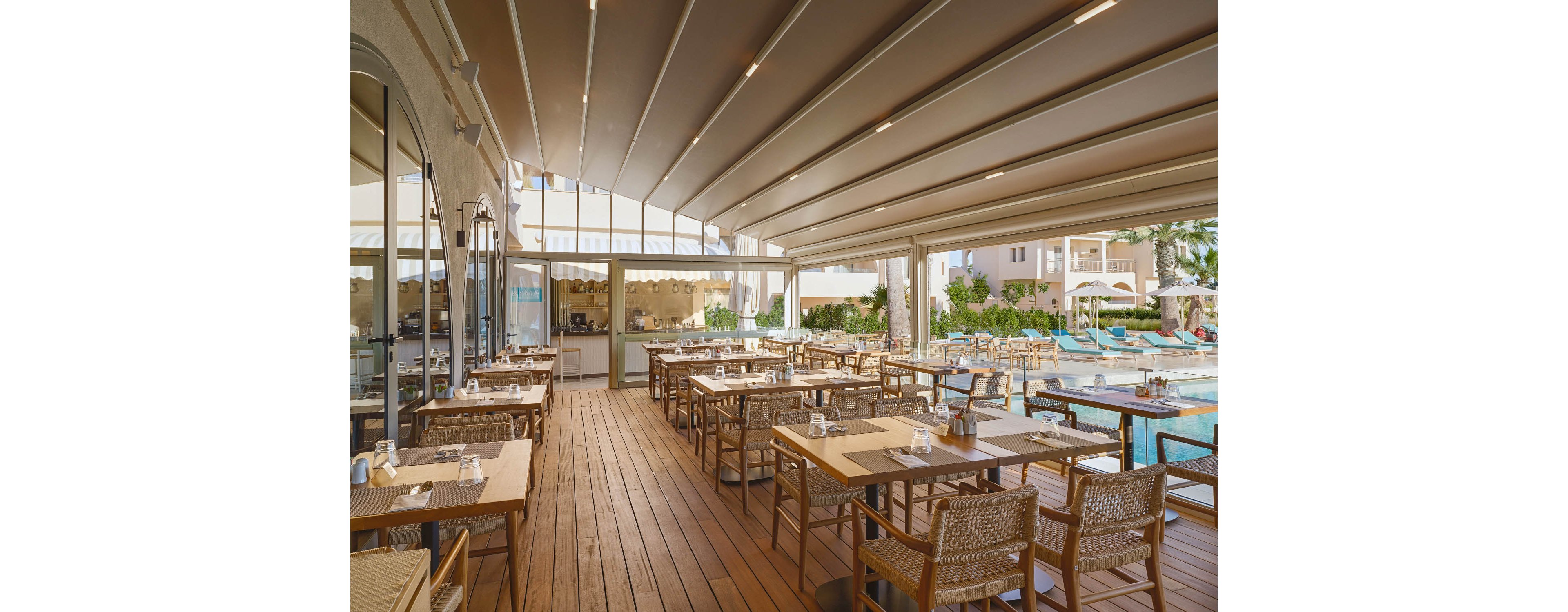 An open-air restaurant features neatly arranged wooden tables and woven chairs on a polished wooden deck. The dining area is covered by a modern retractable roof, allowing natural light to filter through. Large glass windows reveal the indoor bar or service area, while the outdoor pool and sun loungers create a resort-like atmosphere. The scene is calm and inviting, suggesting a relaxed vacation dining experience.
