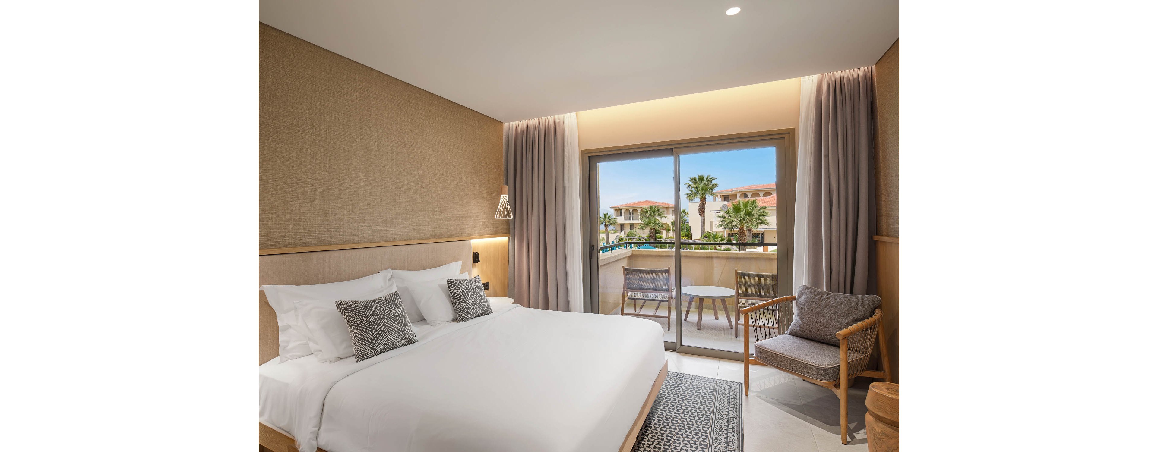 A contemporary hotel bedroom features a large neatly made bed with white linens and patterned accent pillows. Neutral-toned walls and curtains create a calm, minimalist atmosphere. A sliding glass door opens to a balcony with a small round table and two chairs overlooking a resort courtyard with palm trees and low-rise buildings. Soft lighting and simple decor emphasize comfort and relaxation.