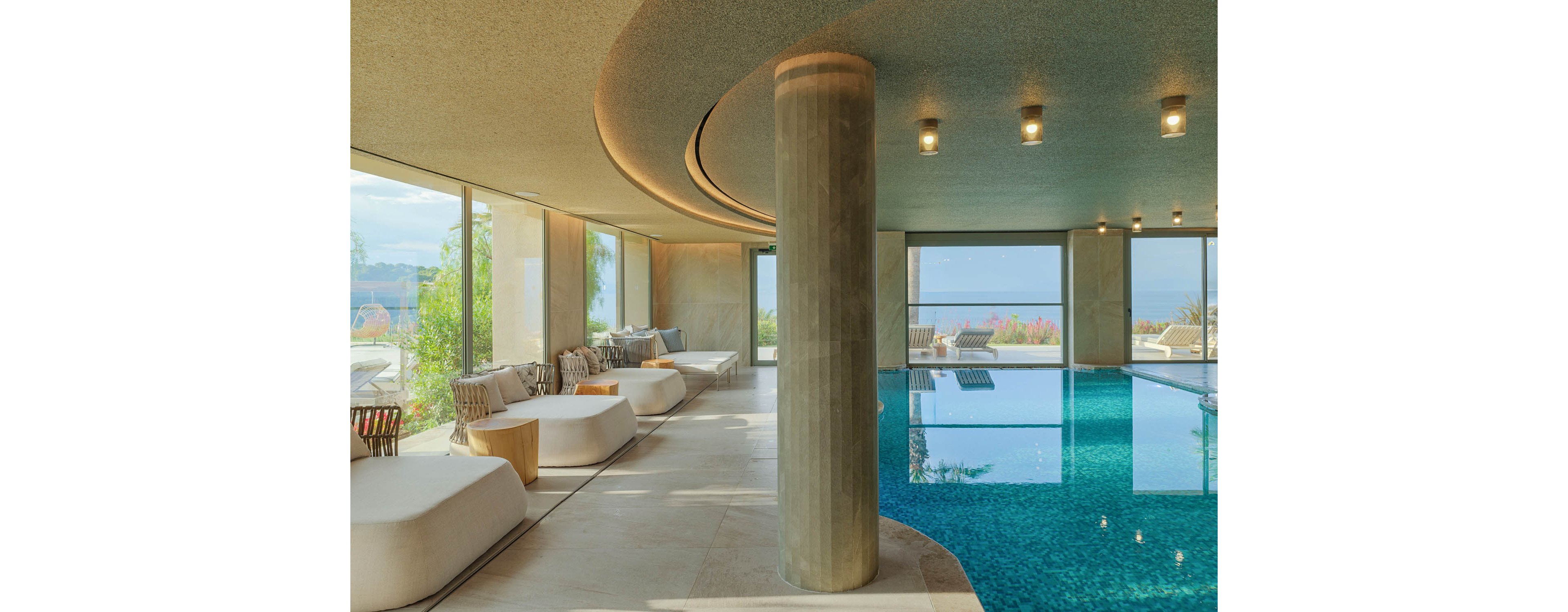 A contemporary indoor swimming pool is set within a spacious lounge featuring large floor-to-ceiling windows. Curved architectural lines, a central concrete column, and recessed ceiling lights create a sleek, modern atmosphere. Cushioned lounge chairs and low tables line the poolside, inviting relaxation. Beyond the glass, greenery and a distant ocean view enhance the tranquil, resort-like setting.