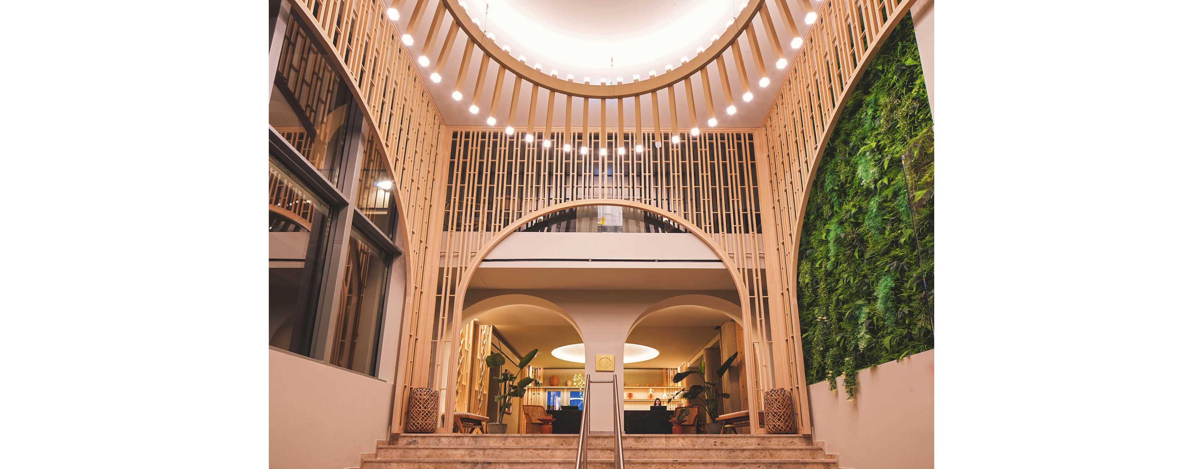 A contemporary interior lobby features tall wooden arches framing a central staircase. Warm lighting from circular ceiling fixtures creates a welcoming atmosphere. On the right side, a lush vertical green wall adds natural texture and color. In the background, seating areas and indoor plants contribute to a stylish, modern design.